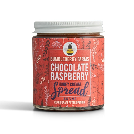 A jar of Bumbleberry Farms Chocolate Raspberry Honey Cream Spread is shown. The clear glass jar has a white lid and a vibrant red label with a pattern of dark leaves and a bee icon. The label specifies the contents as an 8 oz. honey cream spread and includes instructions to refrigerate after opening.