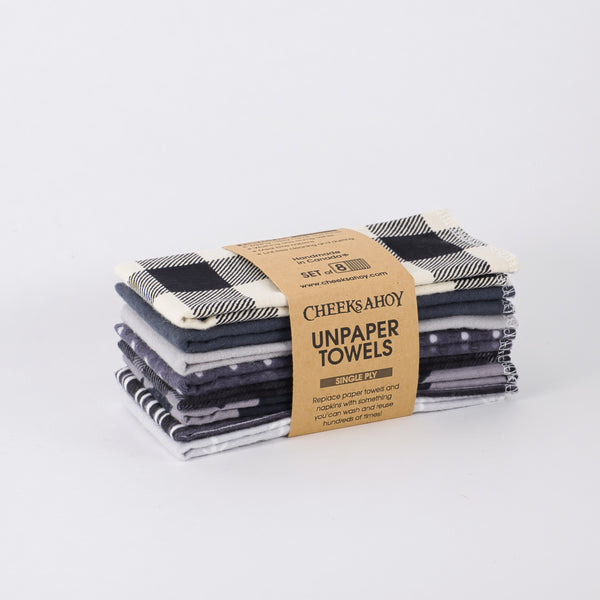 A stack of eight Cheeks Ahoy Unpaper Towels is shown, consisting of various black, white and gray patterns, including a buffalo check print. The cloths are held together by a paper band that identifies them as a set of eight single-ply towels.