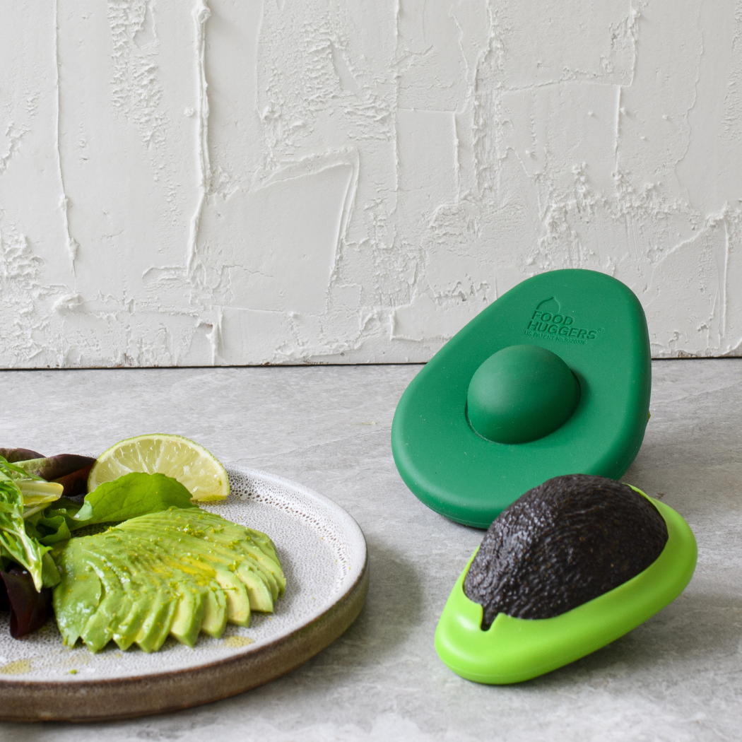 A delightful scene of sustainable food storage, where a large, bright green avocado hugger sits next to a freshly sliced avocado. The other half of the avocado is nestled in a light green hugger, demonstrating how the product can be used to keep food fresh.