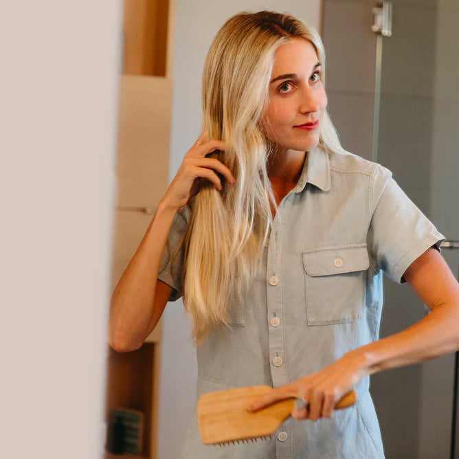 Person brushing long blonde hair with the FTO bamboo hairbrush, shown from behind in a softly lit bathroom setting.