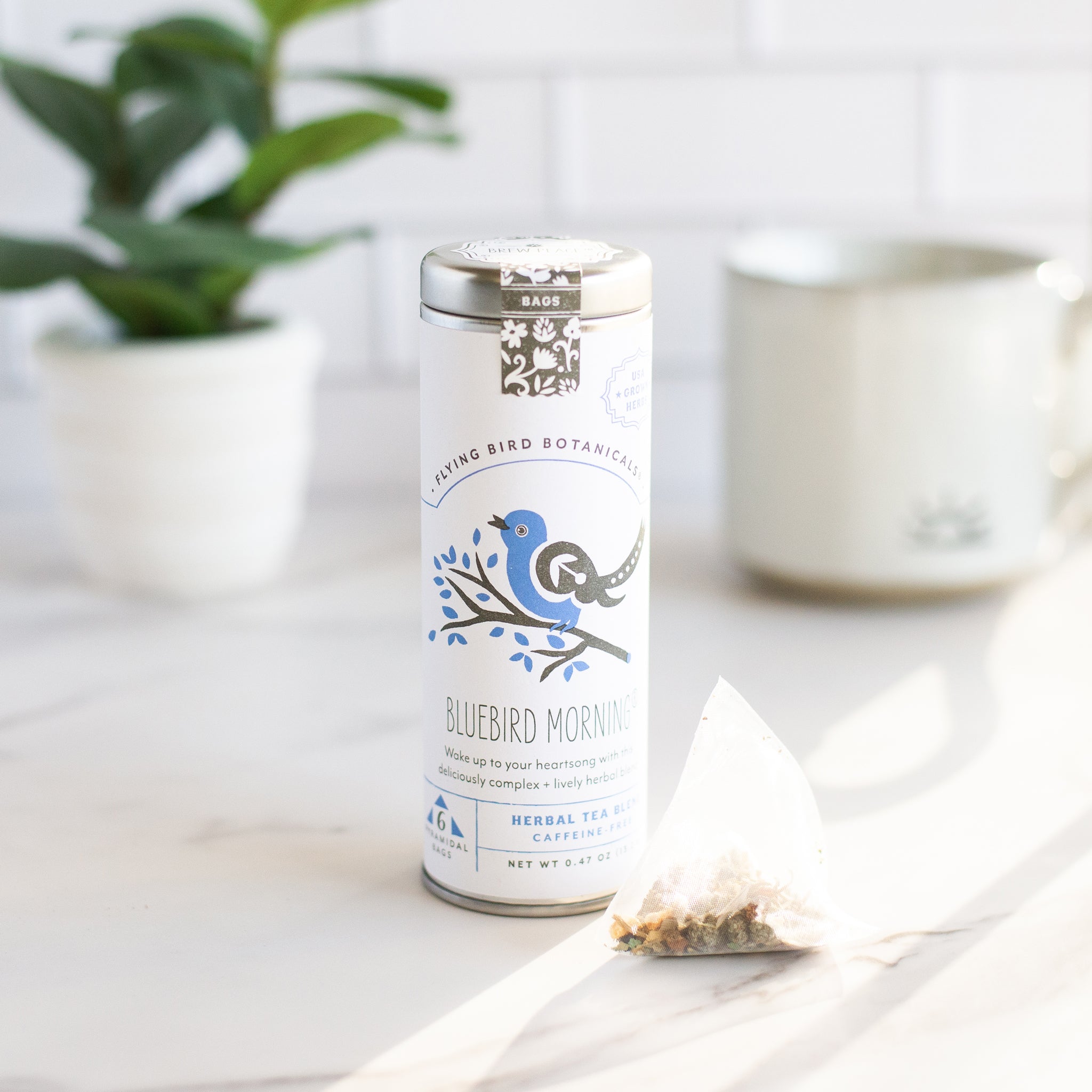 A metal tin of "Bluebird Morning" herbal tea from Flying Bird Botanicals is shown next to a single pyramid shaped tea bag.