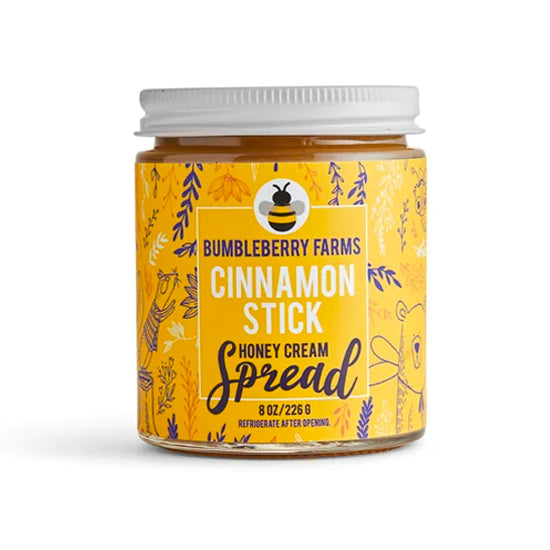 A jar of Bumbleberry Farms Cinnamon Stick Honey Cream Spread is shown. The clear glass jar has a white lid and a bright yellow label with a pattern of dark blue leaves and a bee icon. The label specifies the contents as an 8 oz. honey cream spread and includes instructions to refrigerate after opening.