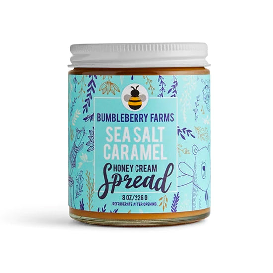 A jar of Bumbleberry Farms Sea Salt Caramel Honey Cream Spread is shown. The clear glass jar has a white lid and a light blue label adorned with a pattern of leaves and a bee icon. The label specifies the contents as an 8 oz. honey cream spread and includes instructions to refrigerate after opening.