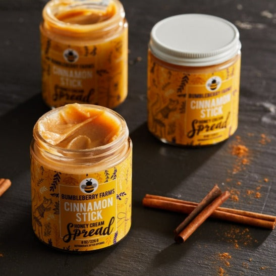 Three jars of Bumbleberry Farms Cinnamon Stick Honey Cream Spread are shown on a dark surface with cinnamon sticks scattered nearby. The jar in the foreground is open, revealing the smooth, golden-brown spread while two other jars are visible in the background.