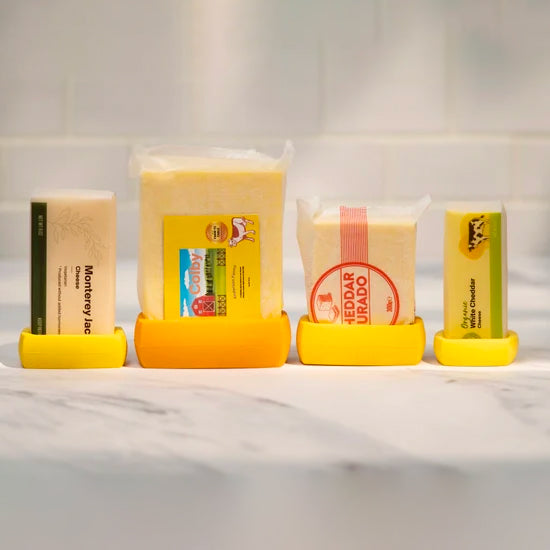Four blocks of cheese are shown on a white marble surface, each contained within a reusable silicone food wrap. The wraps are in varying shades of yellow and orange.