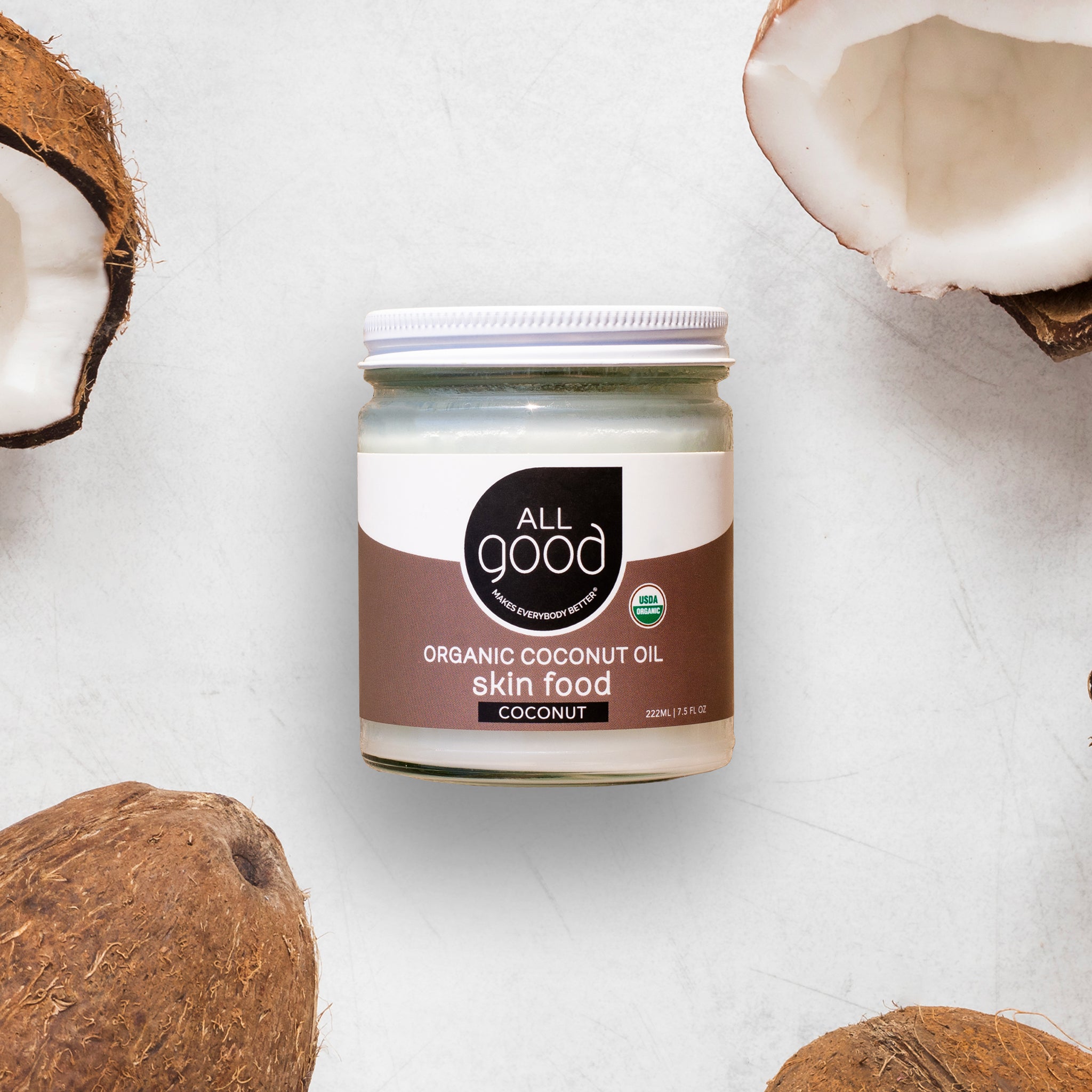 A closed jar of All Good Organic Coconut Oil Skin Food, certified USDA Organic, is surrounded by whole and split coconuts emphasizing its natural origin.
