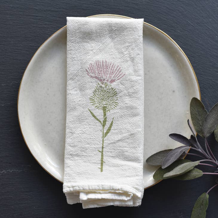 A charming white napkin with a single pink and green thistle design is folded and placed on a white plate with a gold rim, with sprigs of purple sage nearby.