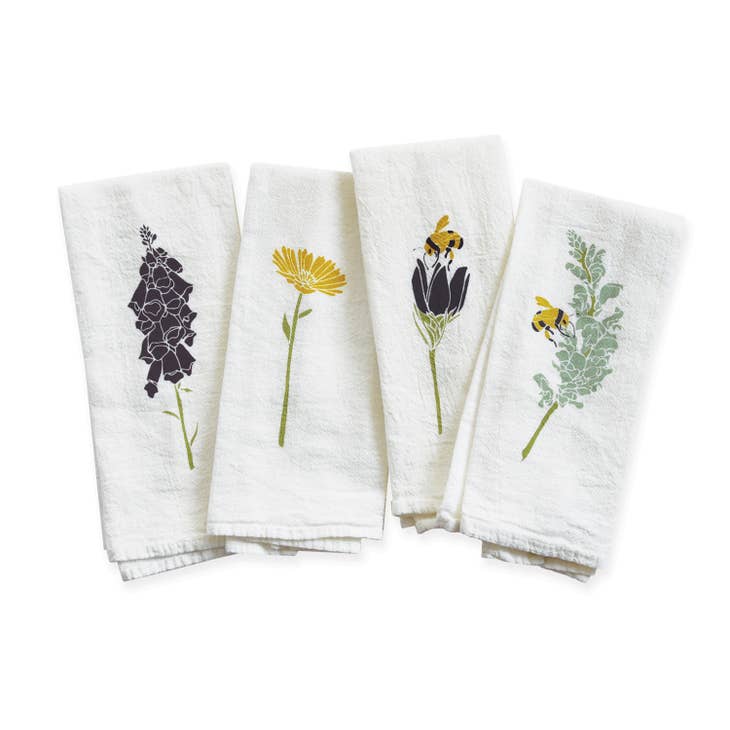 A sweet set of four white napkins is shown folded, each with a different botanical design. The patterns include a dark purple flower, a yellow flower, a purple flower with a busy yellow bee and a green plant with a bee.
