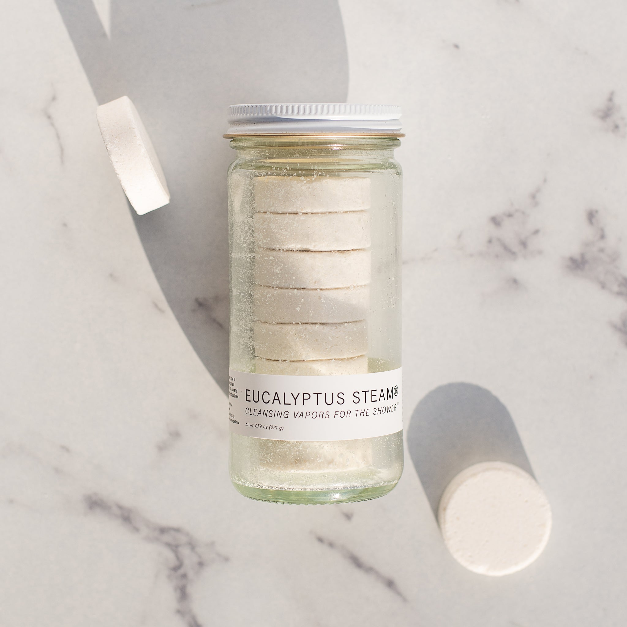 A white lidded glass jar of 'Eucalyptus Steam' cleansing vapors rests on a white marble surface. eight round, white vapors are stacked inside the jar, and two loose vapors are scattered nearby.