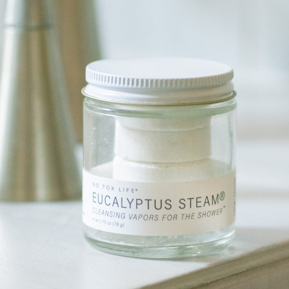 A glass jar from 'No Tox Life' containing a stack of white, round eucalyptus steam tablets. The label reads 'EUCALYPTUS STEAM® Cleansing Vapors for the Shower', weight 2.75 oz (78 g)