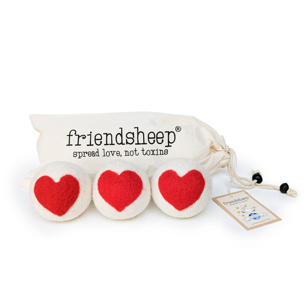 A set of three Friendsheep sustainable goods wool dryer balls are shown, decorated with a cute red heart. They are sitting in front of their white canvas drawstring bag.