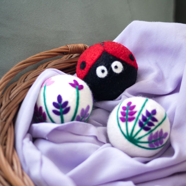 A set of three adorable Friendsheep sustainable goods wool dryer balls, with a ladybug and two purple floral designs, are nestled in a wicker basket on a soft lavender cloth.