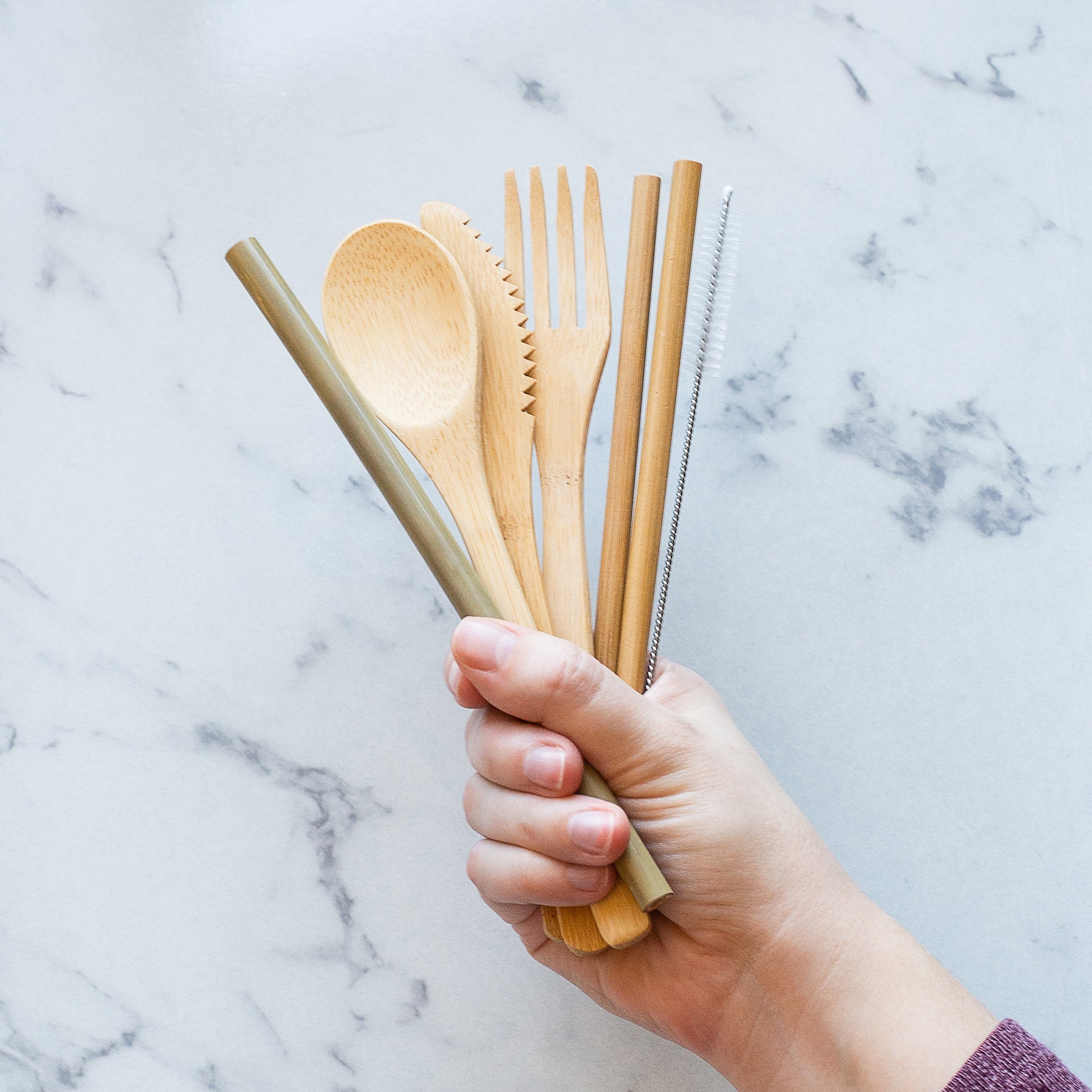 A hand is shown holding a bundle of reusable bamboo utensils against a white marble background. The set includes a spoon, a serrated knife, a fork, chopsticks, a straw and a cleaning brush.