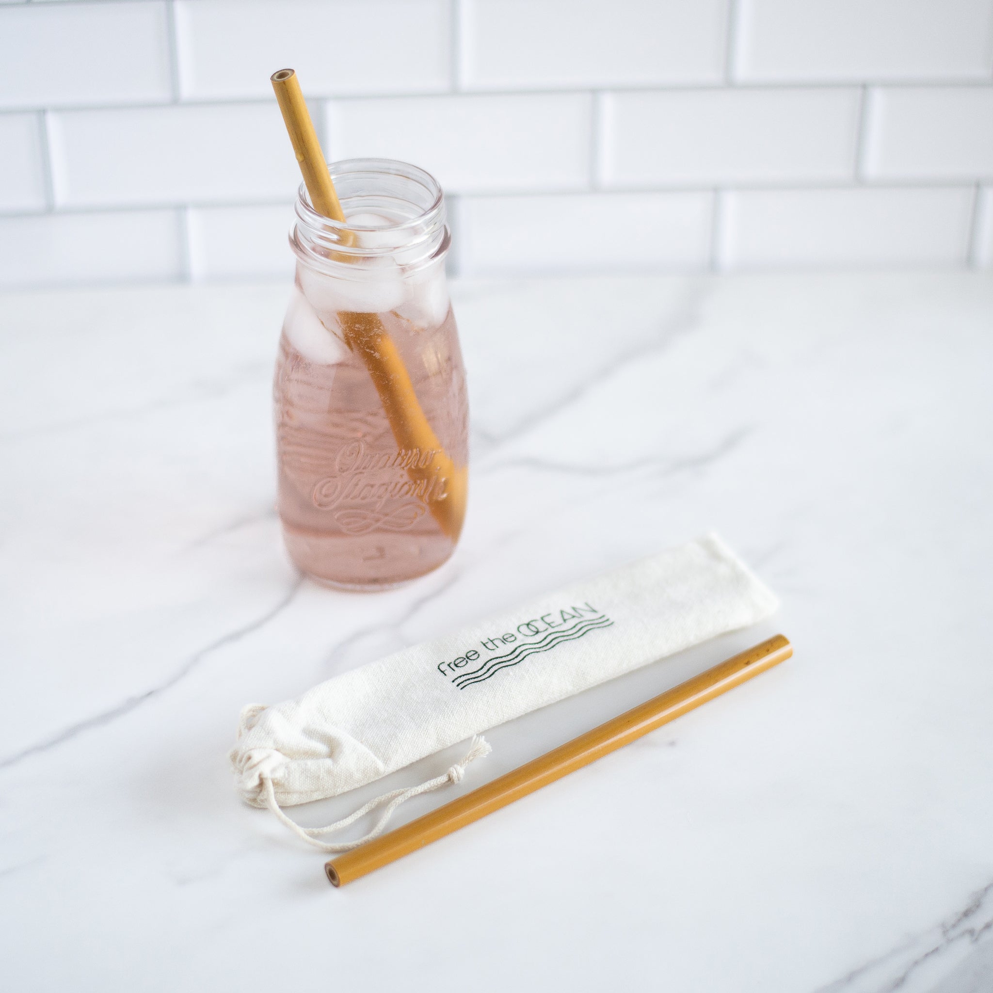 A glass bottle of pink liquid with a bamboo straw and ice cubes sits on a marble countertop. Beside it, a bamboo straw and a cloth carrying bag