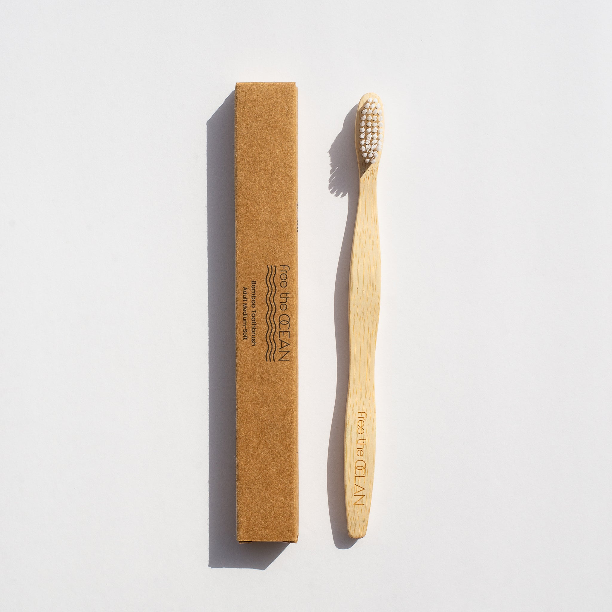 Bamboo toothbrush next to its recyclable brown box, with white bristles and engraved handle.