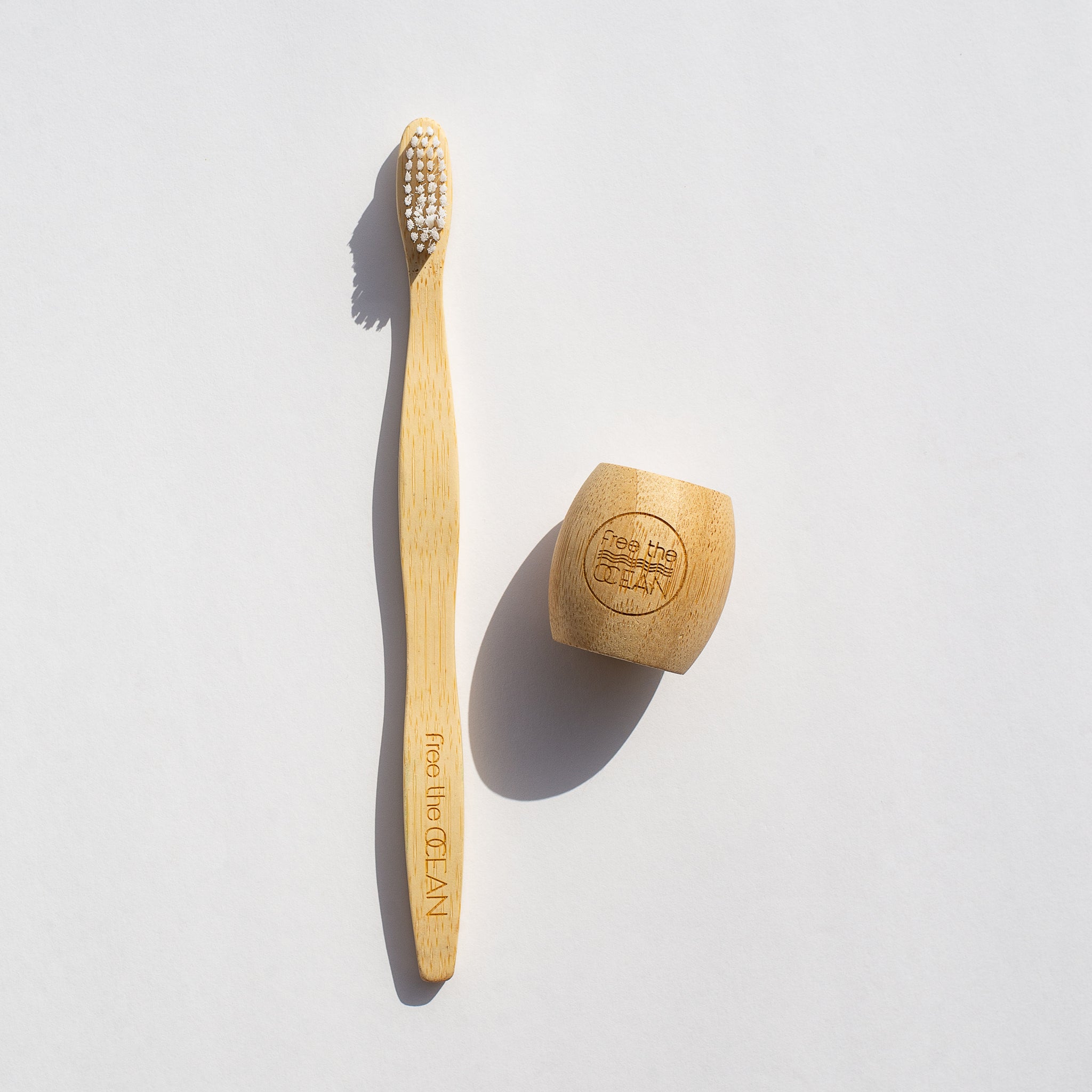 A bamboo toothbrush and a matching wooden holder, both engraved with the "Free the OCEAN" logo are shown on a white surface with a shadow.