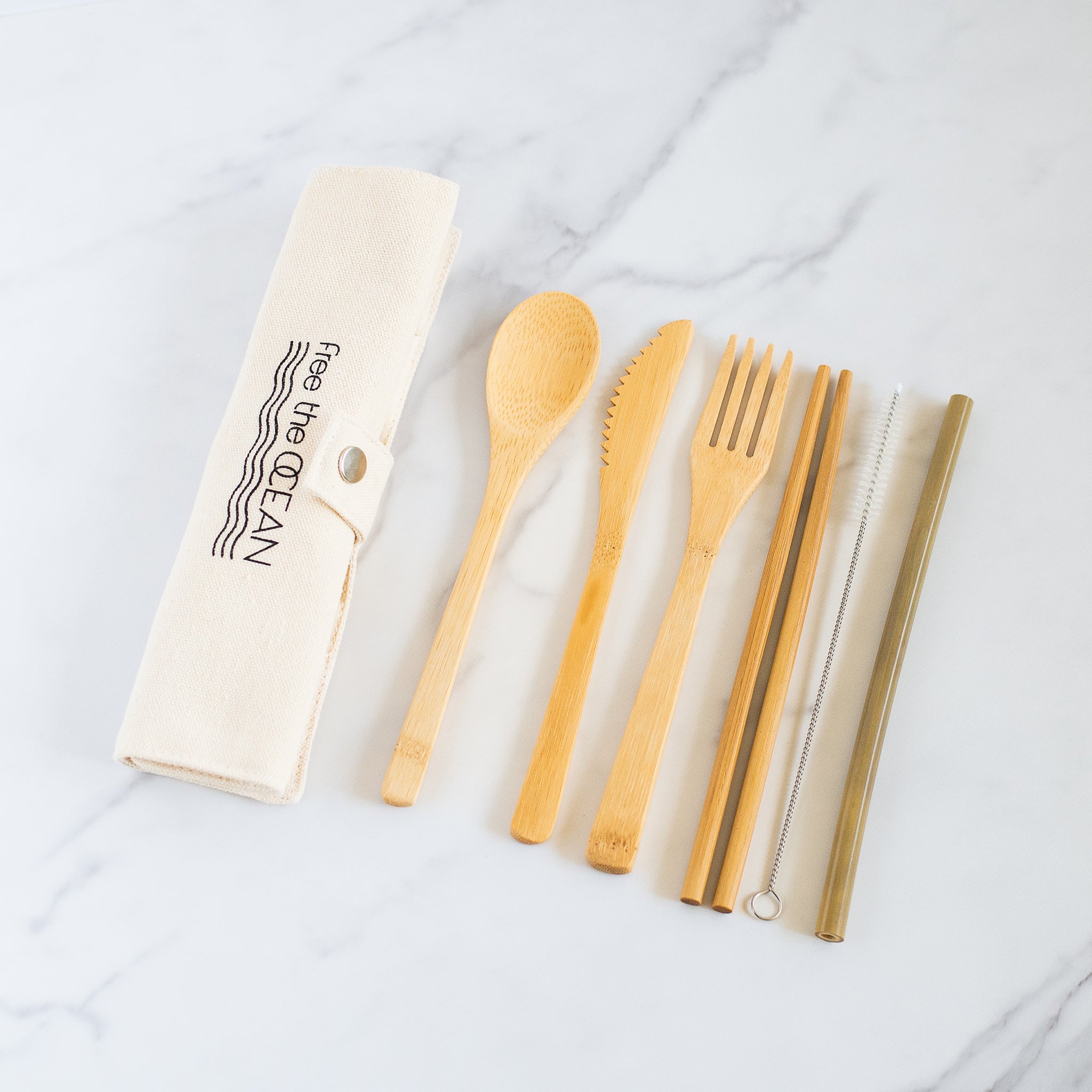 A set of bamboo utensils is shown next to an unrolled beige fabric case that has a Free the OCEAN logo printed on it. The set includes a spoon, a serrated knife, a fork, chopsticks, a straw and a cleaning brush.