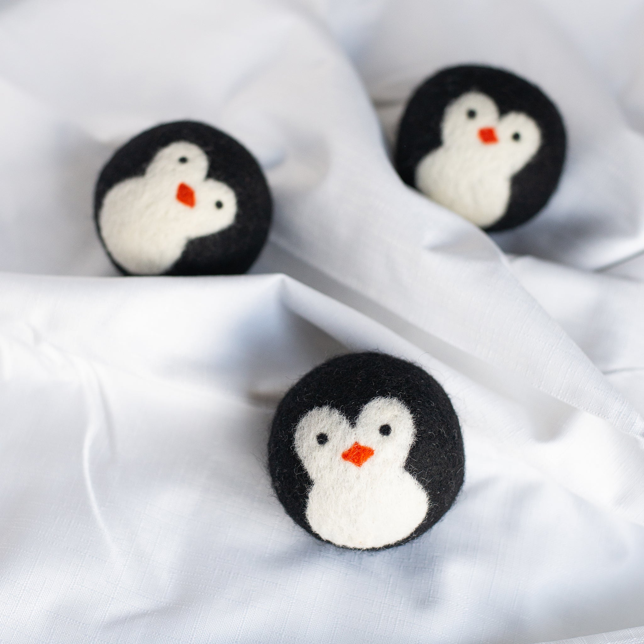 A set of three adorable Friendsheep sustainable goods wool dryer balls, each with a cute penguin design are resting on a rumpled white cloth.