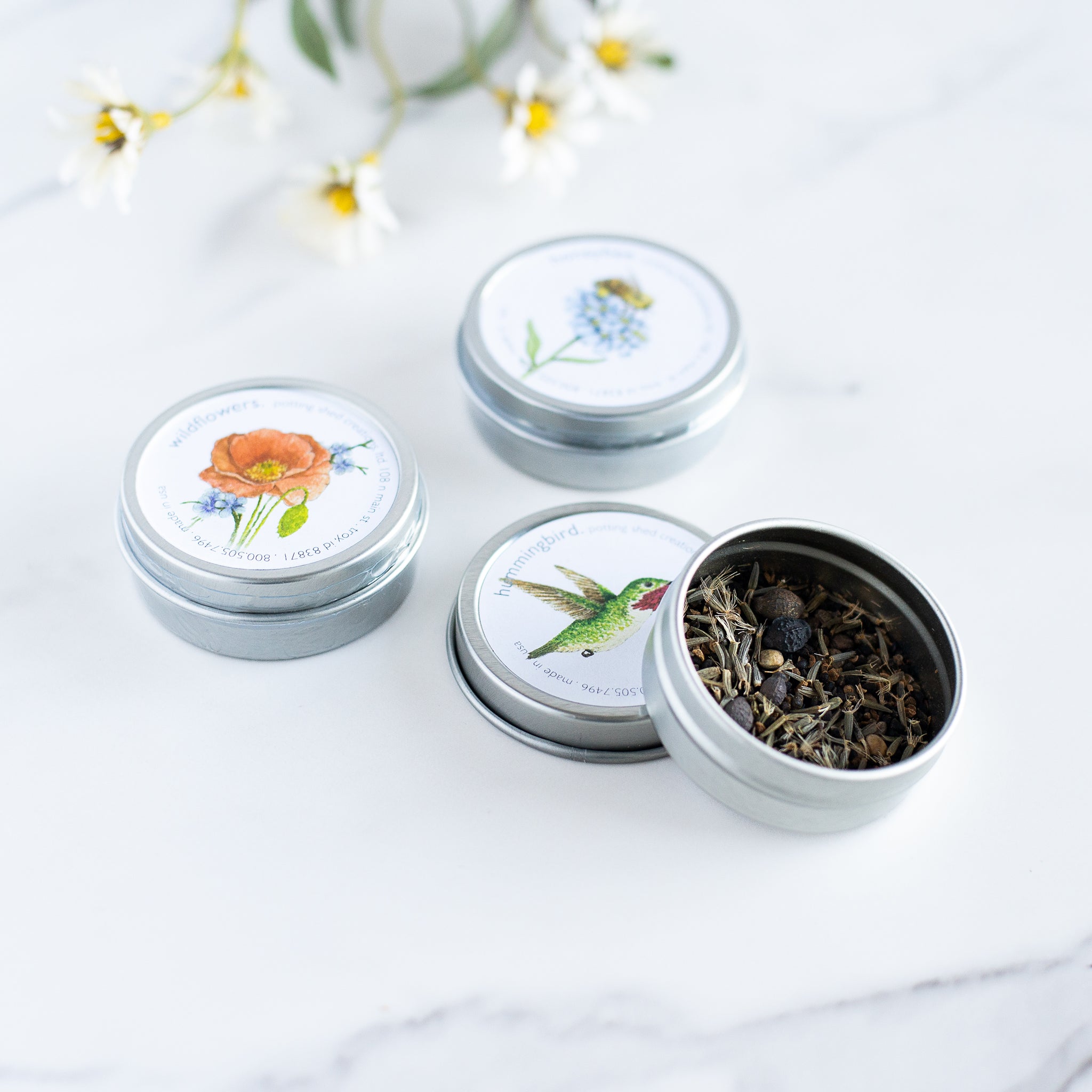 A collection of 3 garden sprinkles tins. The lids feature beautiful illustrations of a poppy, a bee, and a hummingbird. One tin is open, revealing the seeds within.