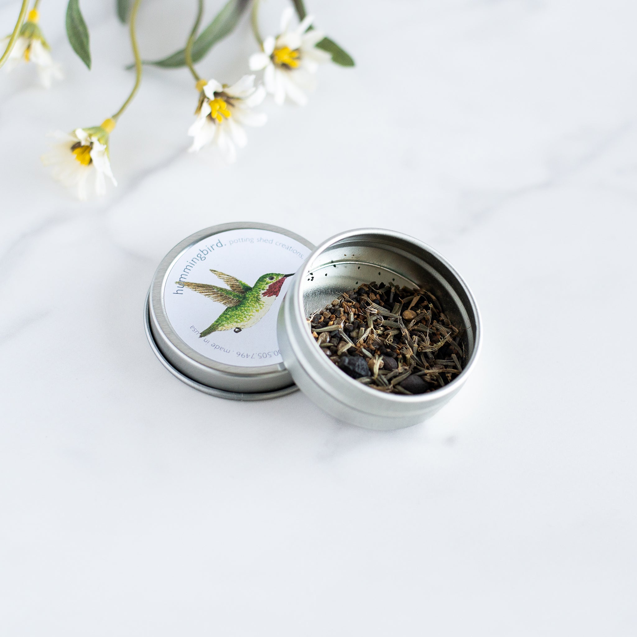 A round, open tin of hummingbird seeds. The lid features a cute illustration of a hummingbird, while the tin itself reveals a mix of dried seeds.