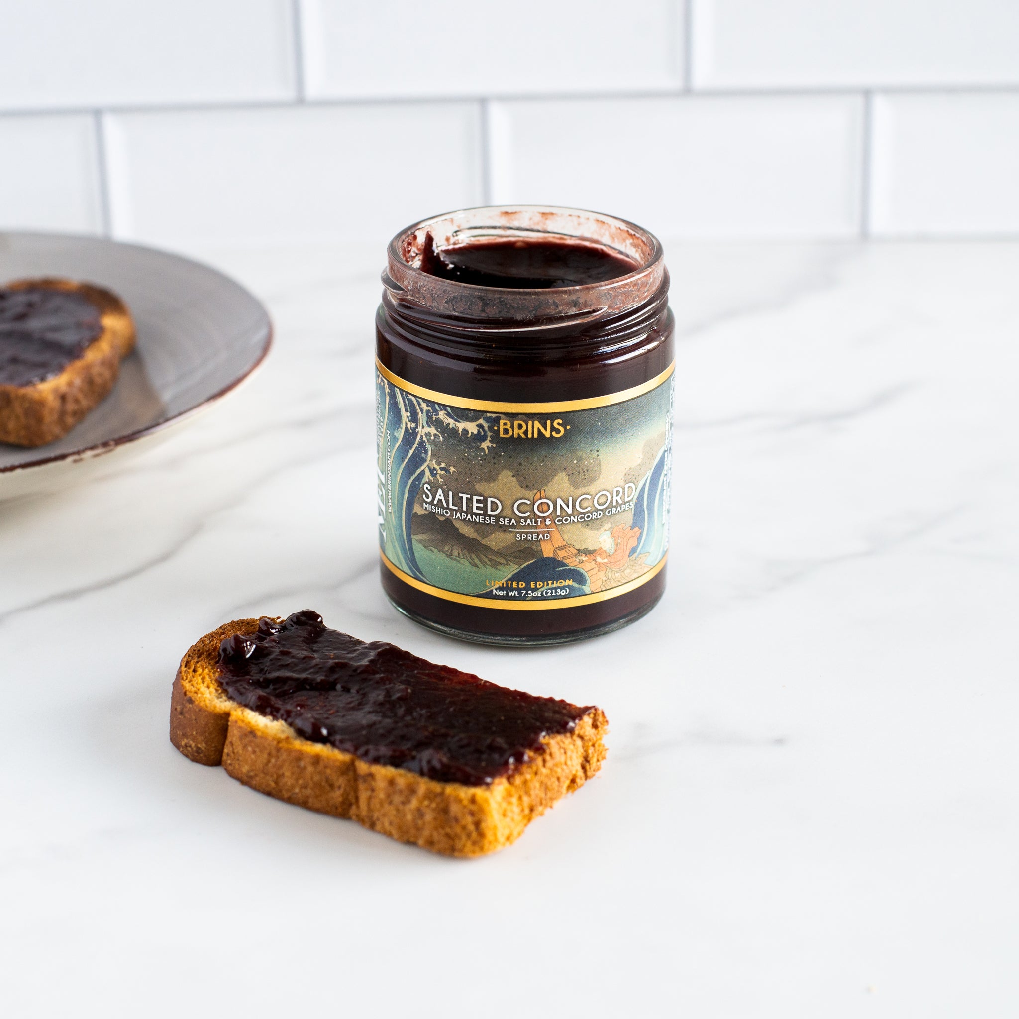 An open jar of Brins Salted Concord spread is shown on a white marble countertop. A piece of toast covered in the dark red spread is next to the jar and another piece is on a plate in the background. The label identifies the spread as containing Mishio Japanese sea salt and Concord grapes.