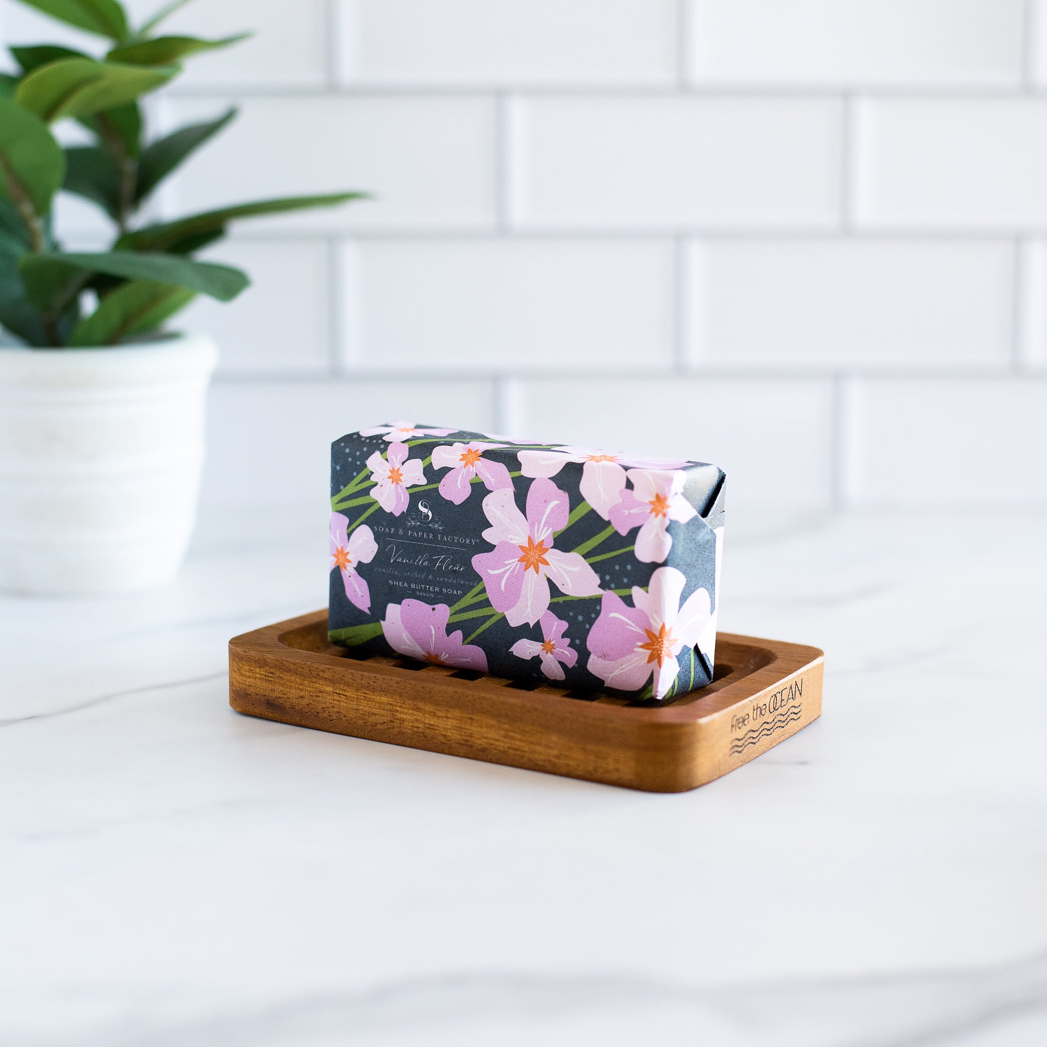 A rectangular "Vanilla Fleur" shea butter soap bar from 'Soap & Paper Factory' sits on a wooden soap dish. The soap dish has "Free the Ocean" carved into the wood.