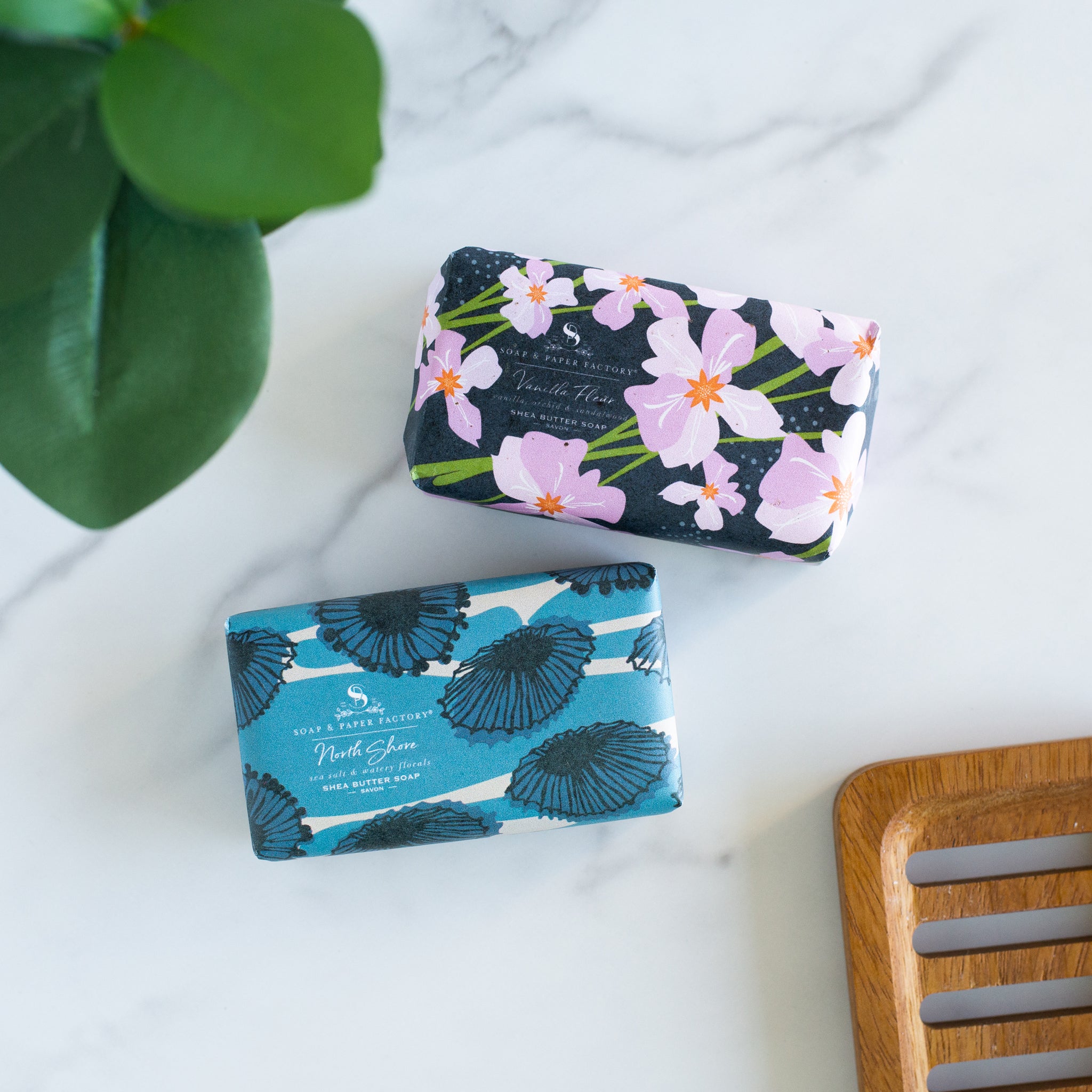 Two 'Soap & Paper Factory' shea butter soaps sit on a marble surface, one wrapped in floral-patterned paper and the other in a blue shell design.