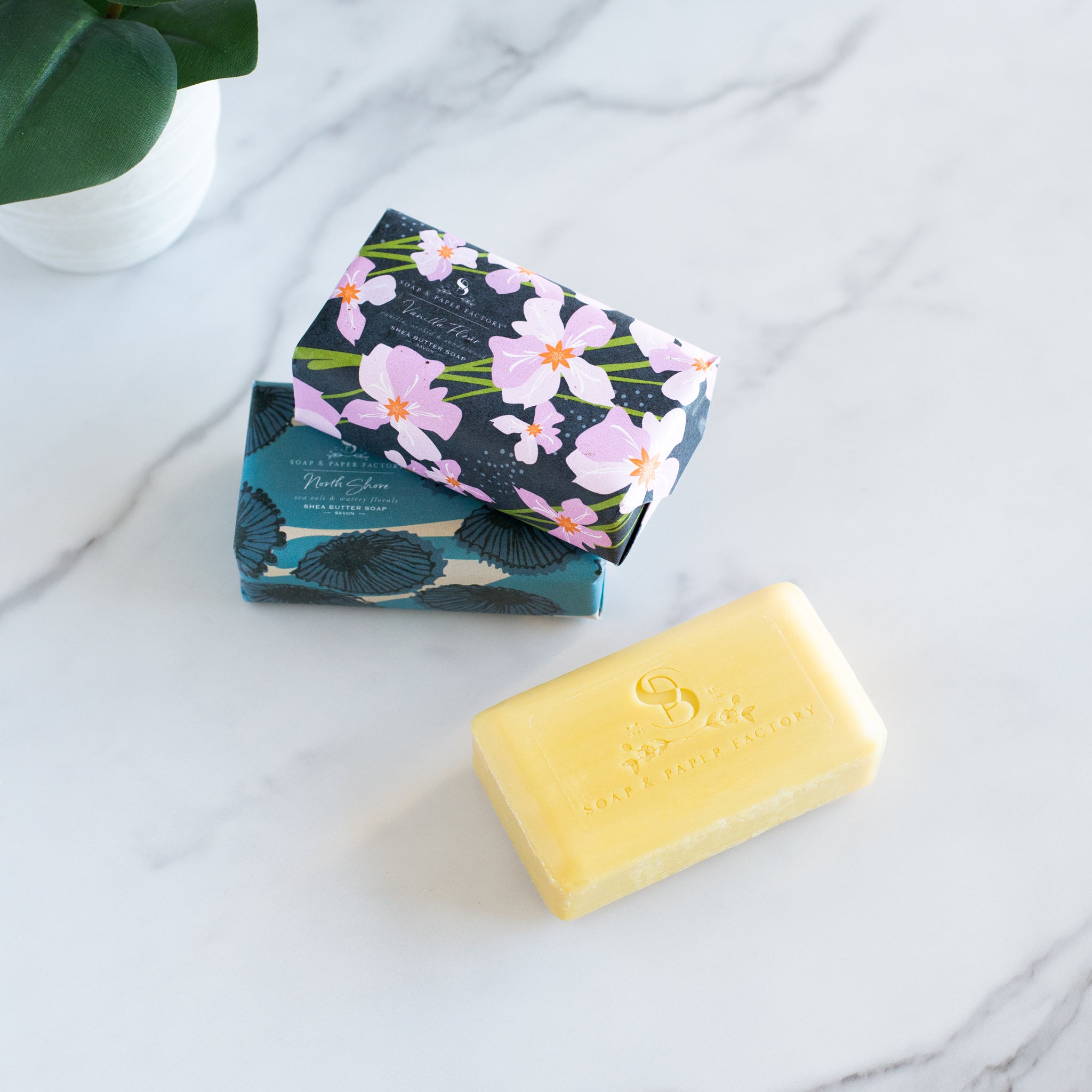 Three Soap & Paper Factory shea butter soaps are on a marble counter, one is an unwrapped yellow bar and two wrapped with floral and shell patterns.
