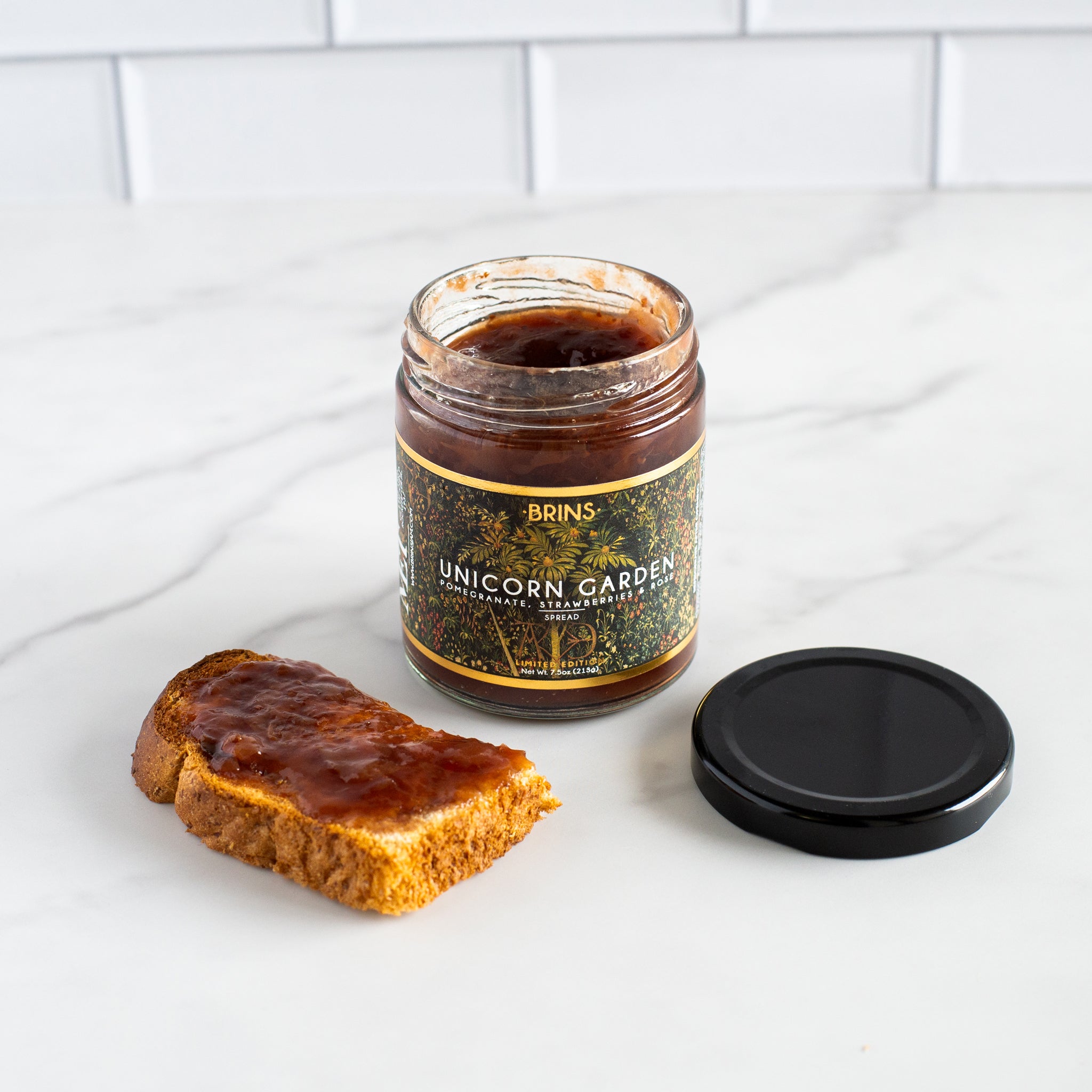 An open jar of Brins Unicorn Garden spread is shown on a white marble countertop with a piece of toast that has been covered in the spread. The jar's black lid is placed next to it. The product is a pomegranate, strawberry and rose-flavored spread.