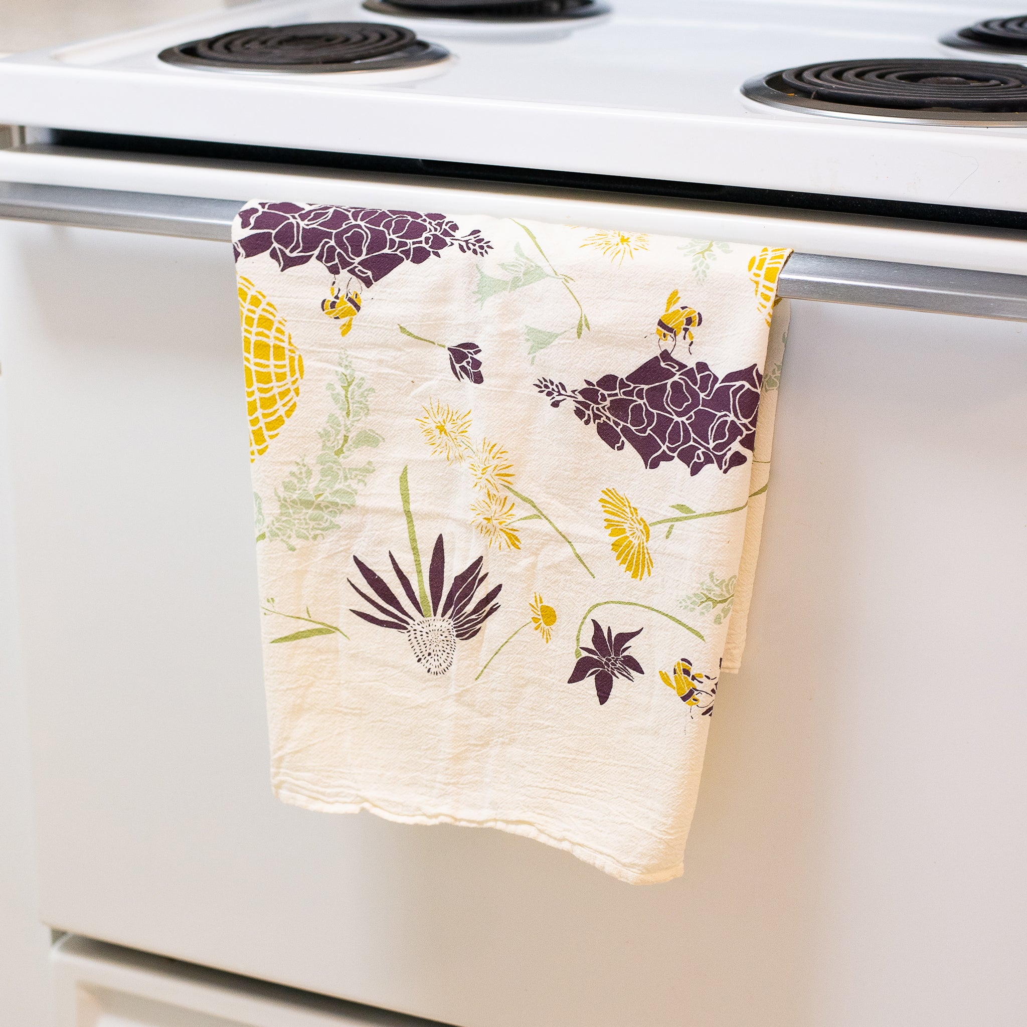 A cheerful white kitchen towel with a playful pattern of yellow bees, beehives and dark purple flowers is hanging from the handle of a white stove.