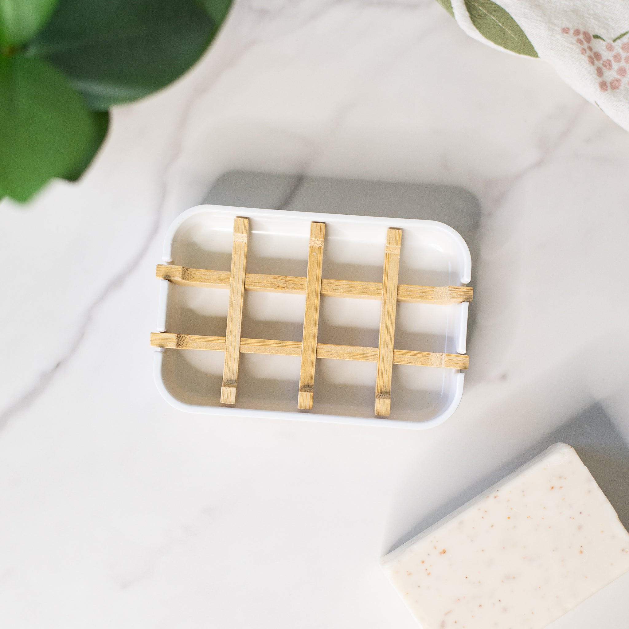 A white soap dish with a bamboo slatted insert sits on a white marble countertop, next to a soap bar.