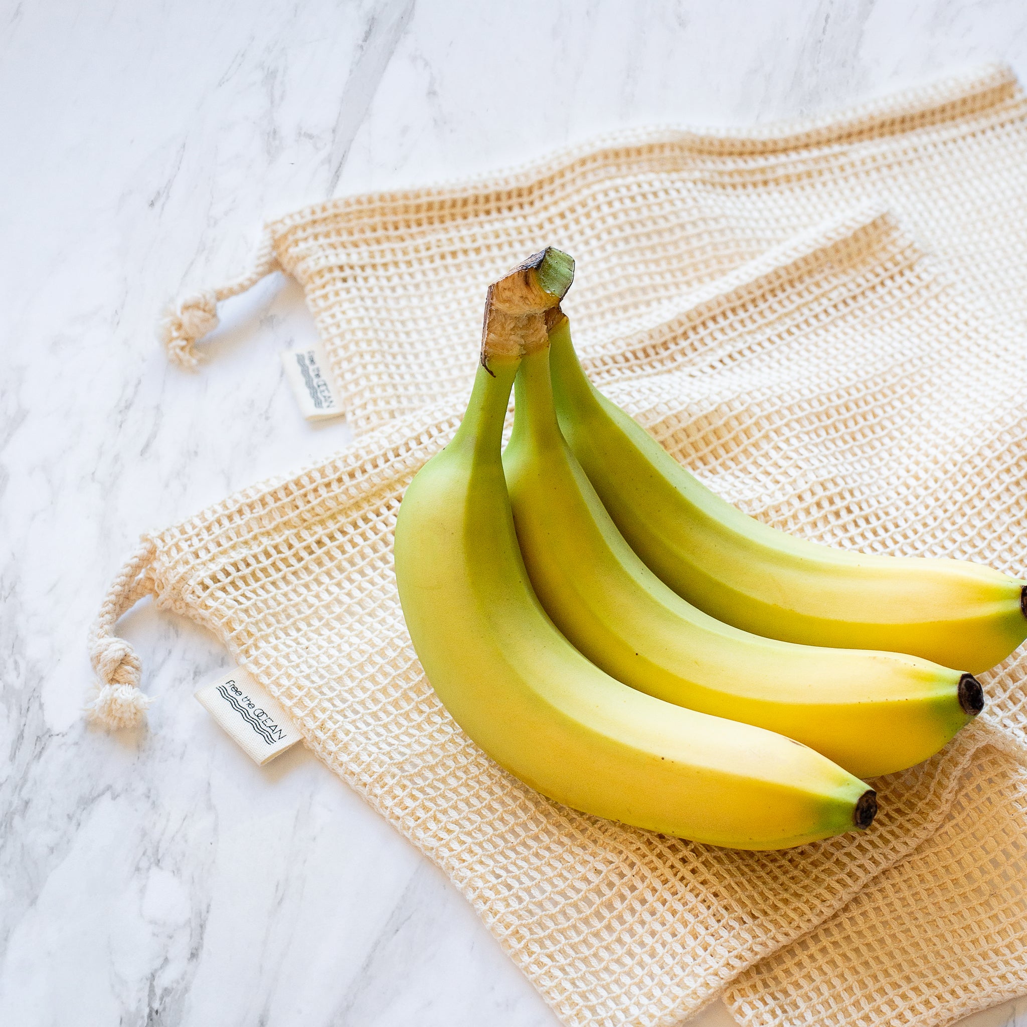 A bunch of three bananas is shown resting on top of two reusable produce bags on a white marble countertop. The bags are made of a natural-colored mesh material and have small tags with text on them.