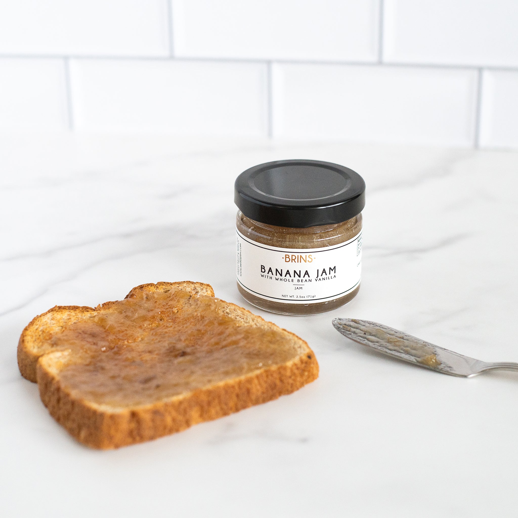 A small jar of "BRINS" Banana Jam sits next to a piece of golden brown toast with a thin layer of the sweet golden jam.