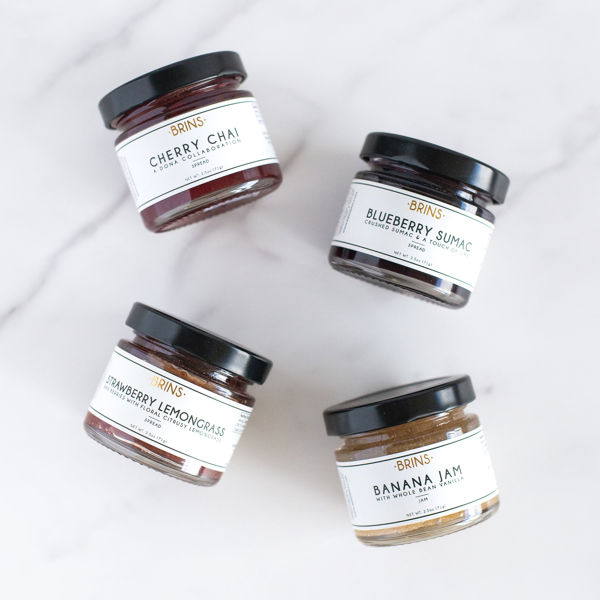 An overhead shot of four small jars of "BRINS" jam on marble: red cherry chai, dark blue blueberry sumac, light red strawberry lemongrass, and tan banana vanilla.