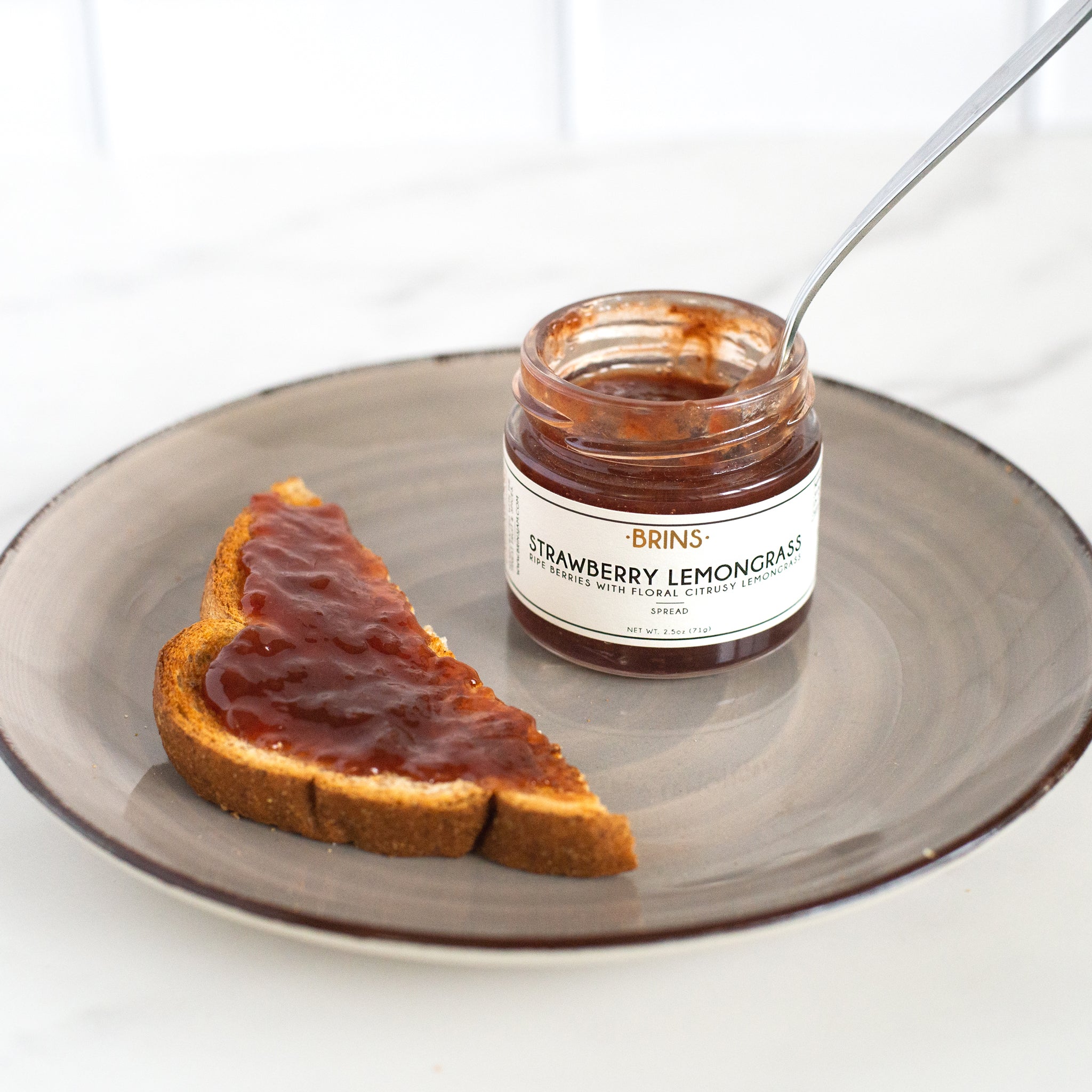 A slice of toast with strawberry lemongrass jam is on a gray plate next to an open jar of the jam with a spoon inside.