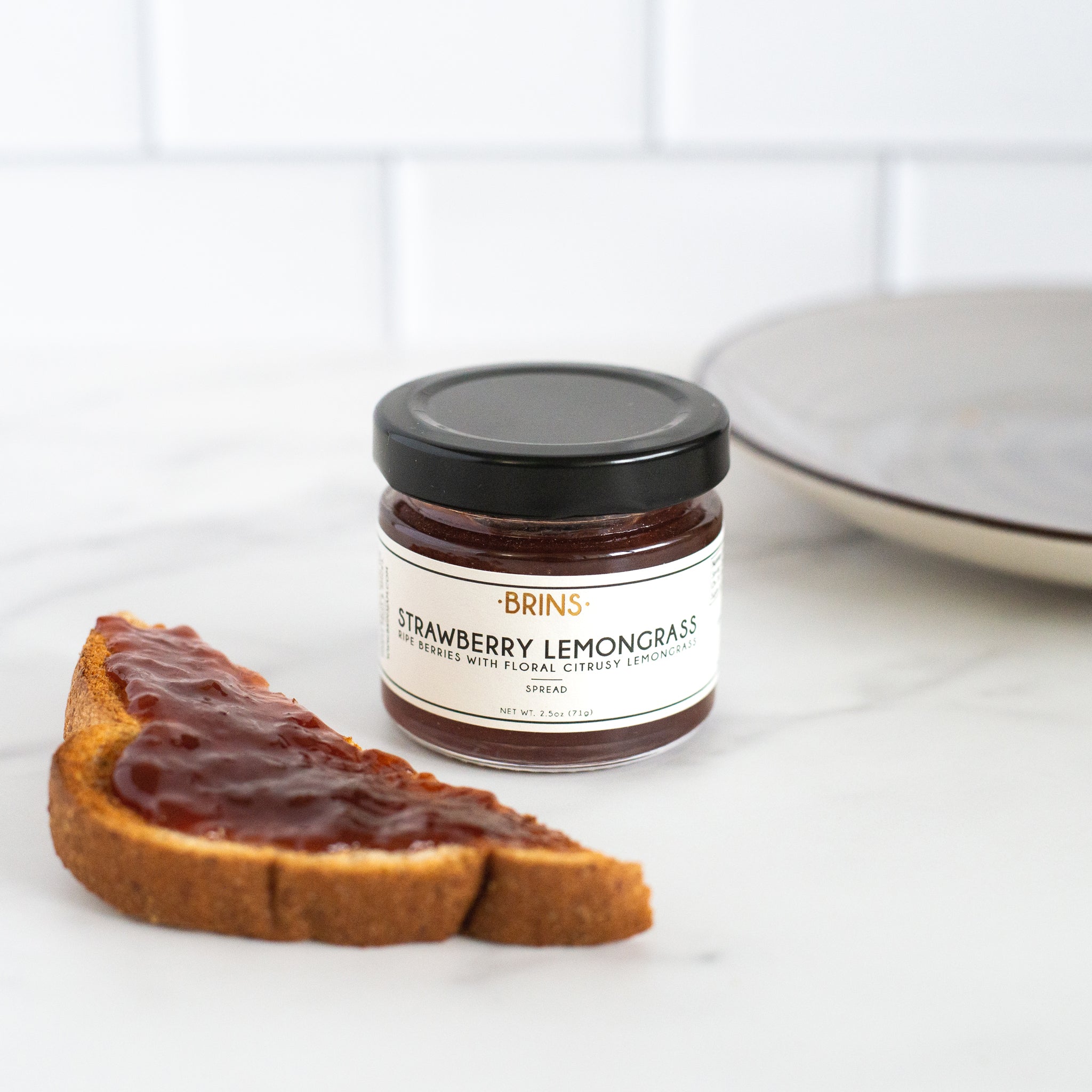 A small jar of "BRINS" Strawberry Lemongrass spread sits next to a piece of golden brown toast with a generous layer of the tasty red jam.
