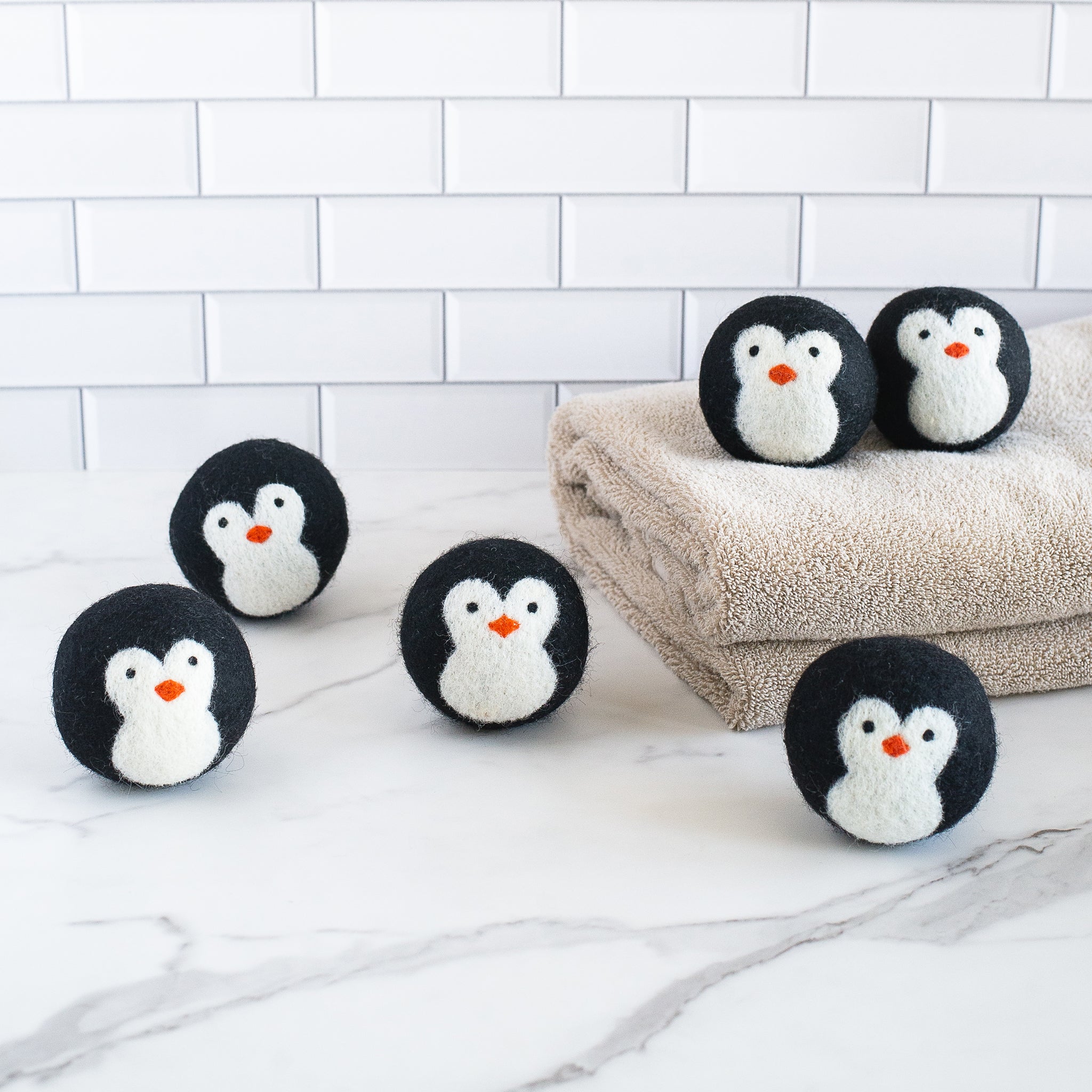 Six Penguin wool dryer balls, two resting on a white towel and four on a marble surface, showing soft textures and black-and-white penguin designs.