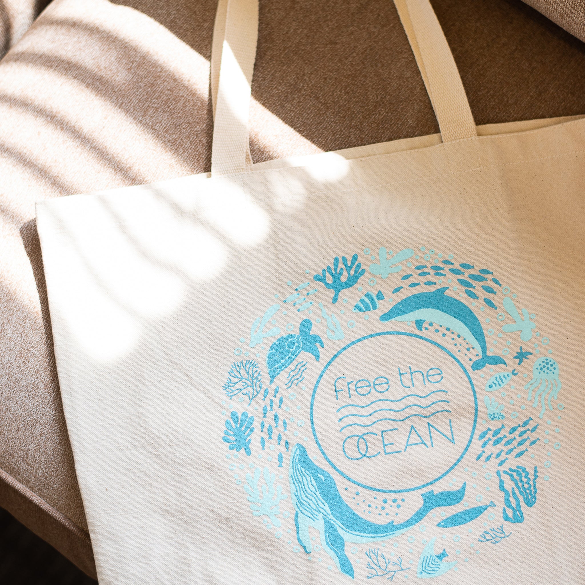 A beige canvas tote bag with a teal-blue logo is resting on a textured couch. The logo features a circle of various sea creatures, with the text "Free the OCEAN" in the center. Sunlight is casting a shadow on the bag.