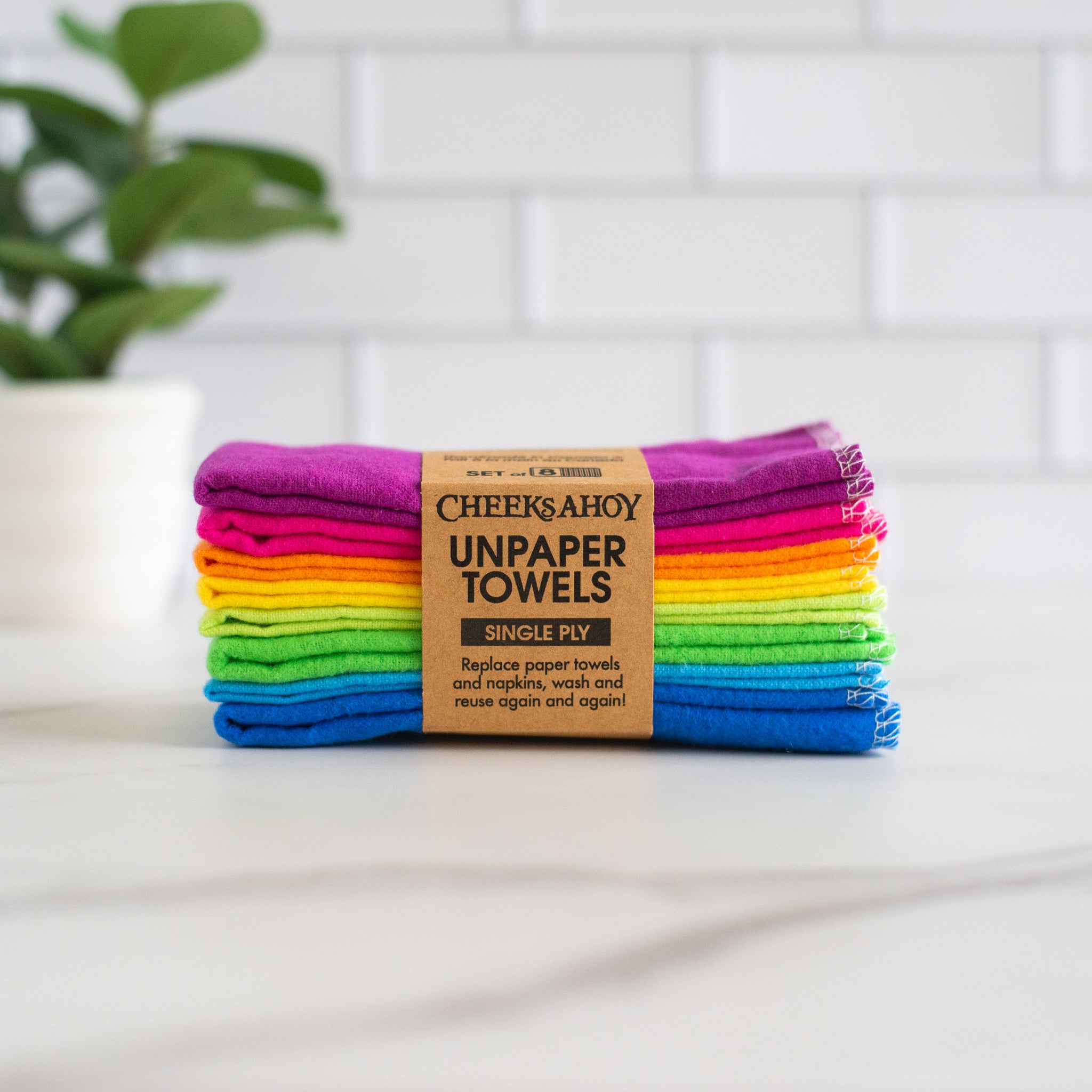 A stack of eight Cheeks Ahoy Unpaper Towels in a rainbow of colors is shown on a countertop. The cloths are held together by a paper band that identifies them as a set of eight single-ply towels.