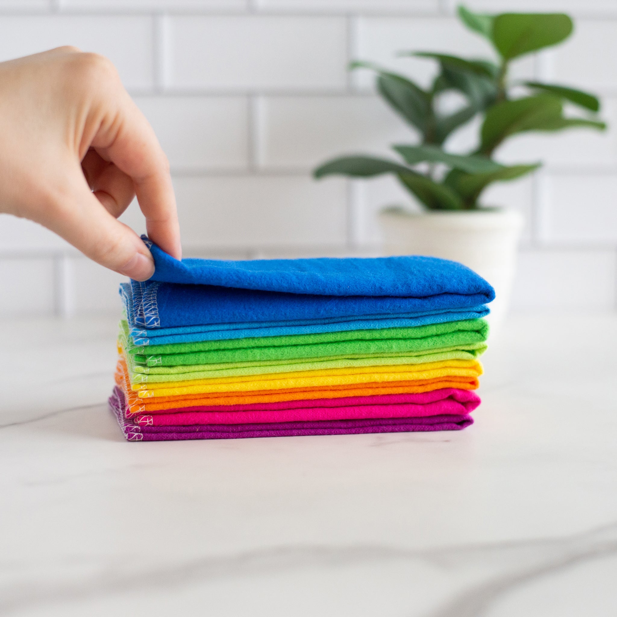 A hand is shown pulling a blue towel from a stack of brightly colored Cheeks Ahoy reusable towels. The towels are in a rainbow color pattern and are sitting on a white marble countertop