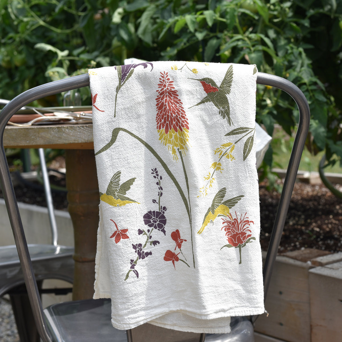 A white tea towel with a charming pattern of hummingbirds and flowers in shades of yellow, red and purple is draped over a metal chair, with lush green plants in the background.