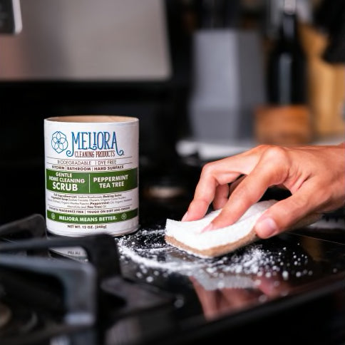A person's hand is shown using a sponge to scrub a black stovetop with a white powder. In the background, a container of Mellora Gentle Home Cleaning Scrub is visible, identifying the powder. The scrub is identified as a "peppermint tea tree" scent.