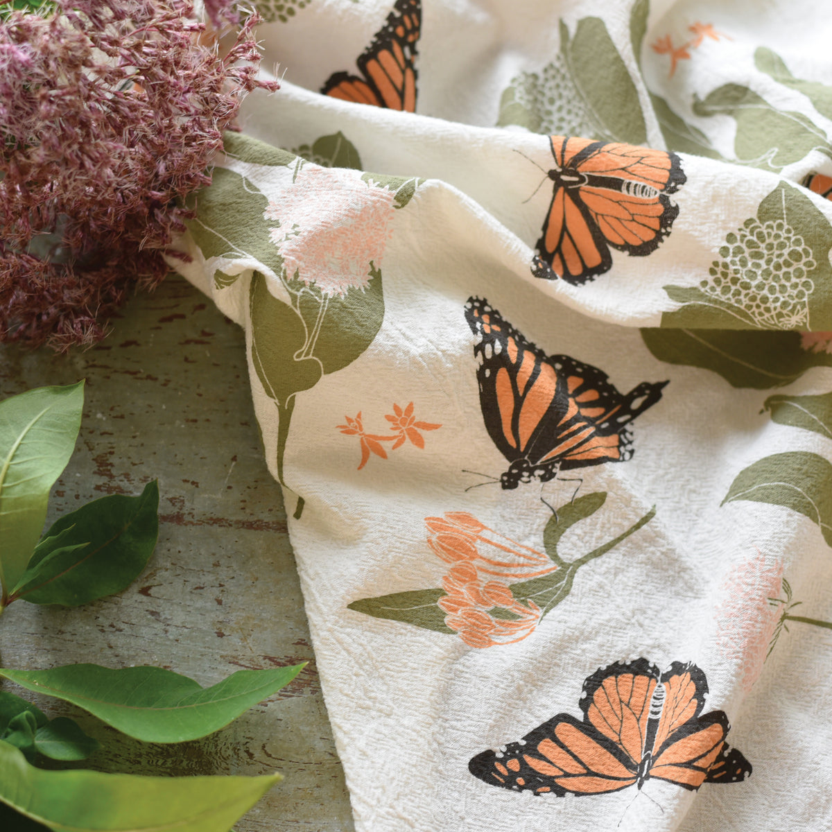 A charming white cotton kitchen towel with a playful print of monarch butterflies and peachy blossoms is draped next to a fuzzy purple allium flower and green leaves on a rustic background.
