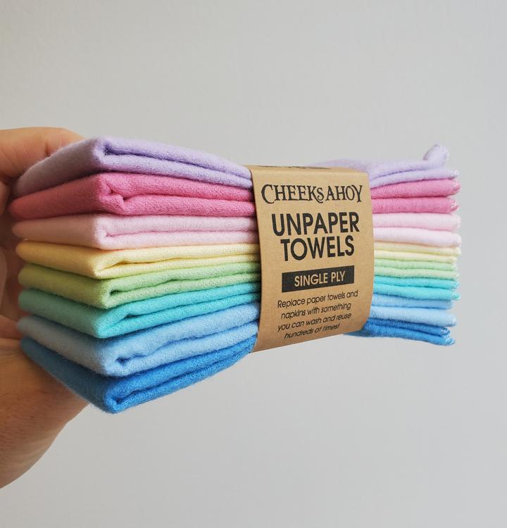 A hand is shown holding a stack of eight Cheeks Ahoy Unpaper Towels  in various pastel shades of the rainbow. The cloths are held together by a paper band that identifies them as a set of eight single-ply towels. The band also states they can be washed and reused hundreds of times to replace paper towels and napkins.