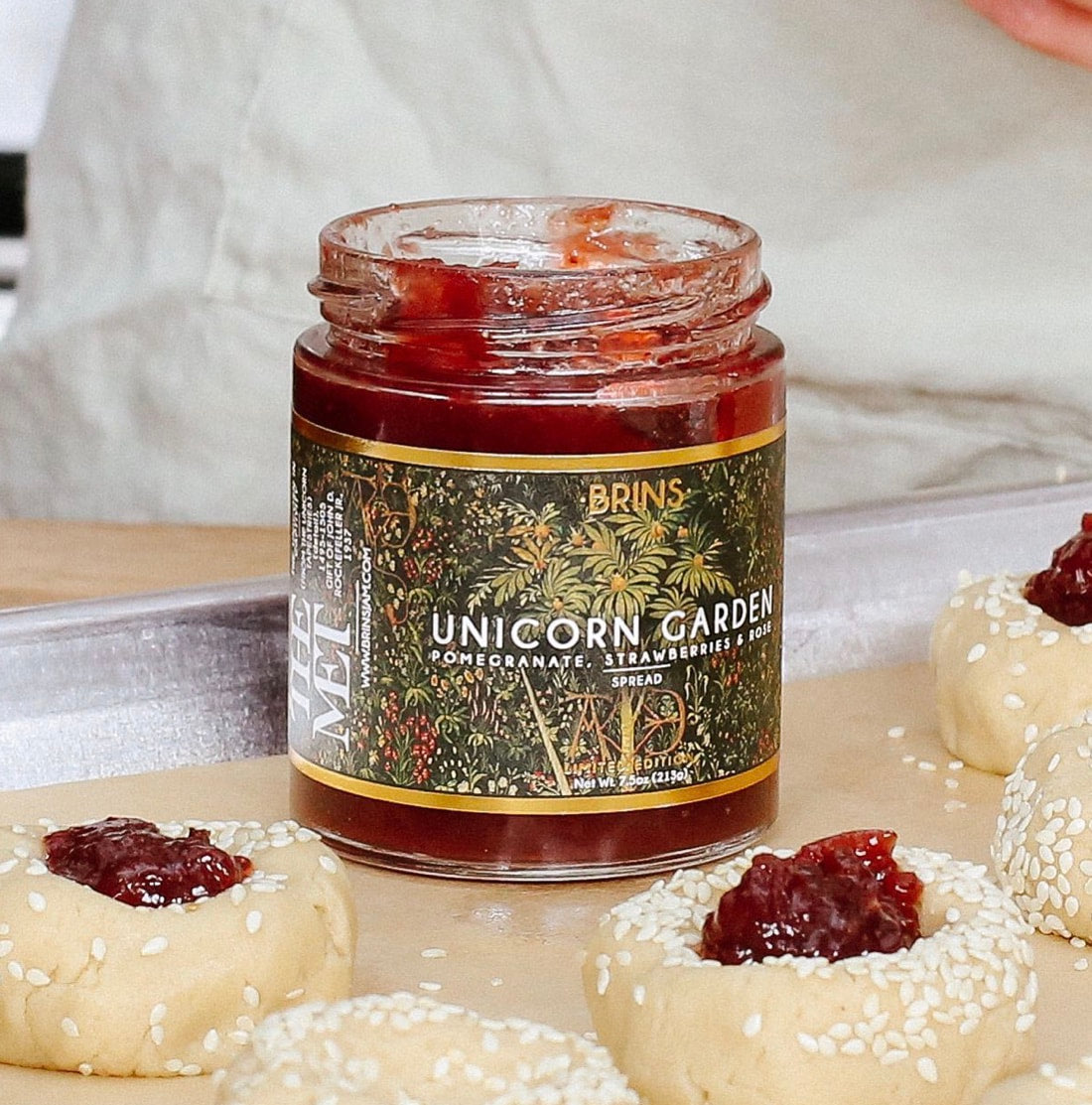 An open jar of Brins Unicorn Garden spread is shown on a baking sheet. Several pieces of dough with sesame seeds are arranged around the jar, each topped with a small amount of the red spread. The product is a pomegranate, strawberry and rose-flavored spread.