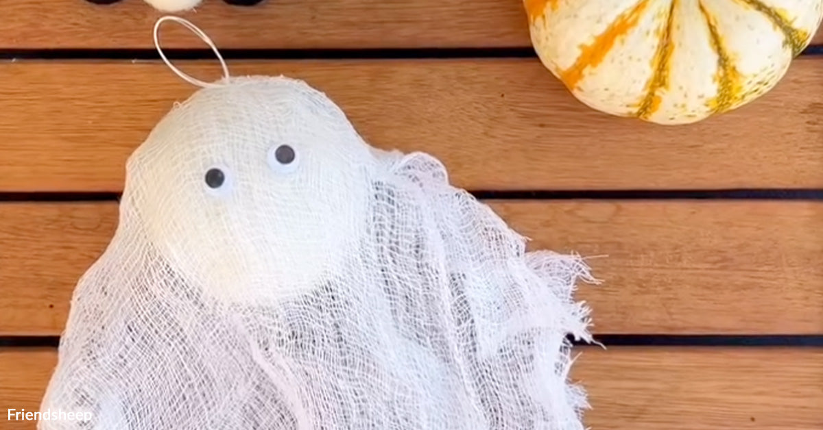 DIY Halloween ghost decoration made from white cheesecloth draped over a round dryer ball with googly eyes, placed on a wooden table beside a small striped pumpkin.