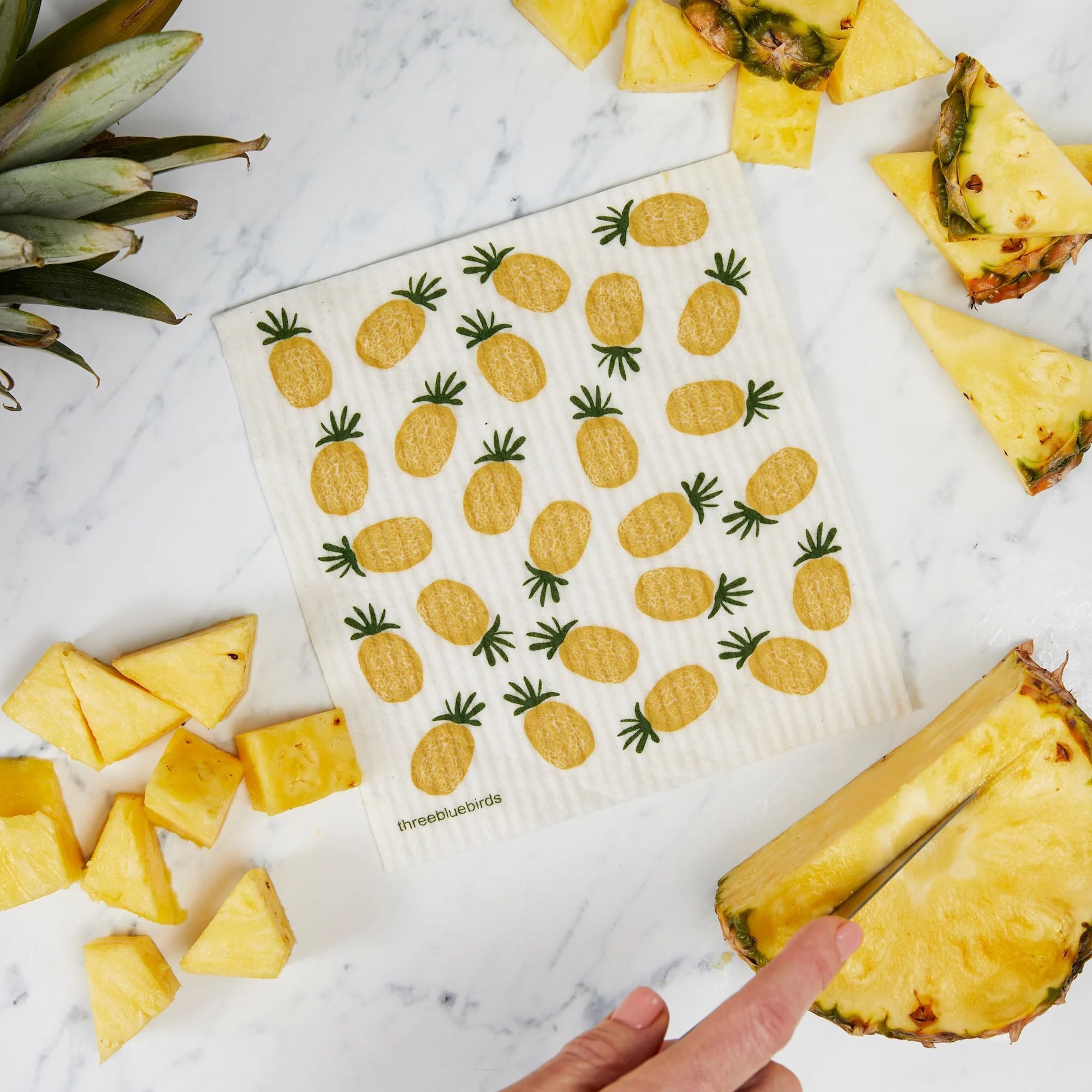 A lifestyle shot showcasing a reusable Swedish dishcloth with a sunny yellow pineapple pattern in a bright kitchen setting cutting pineapples
