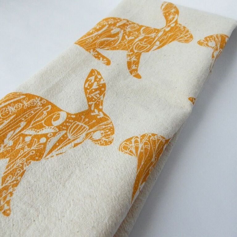 A hand-printed organic cotton tea towel, features a repeating pattern of stylized orange rabbits filled with intricate floral designs.