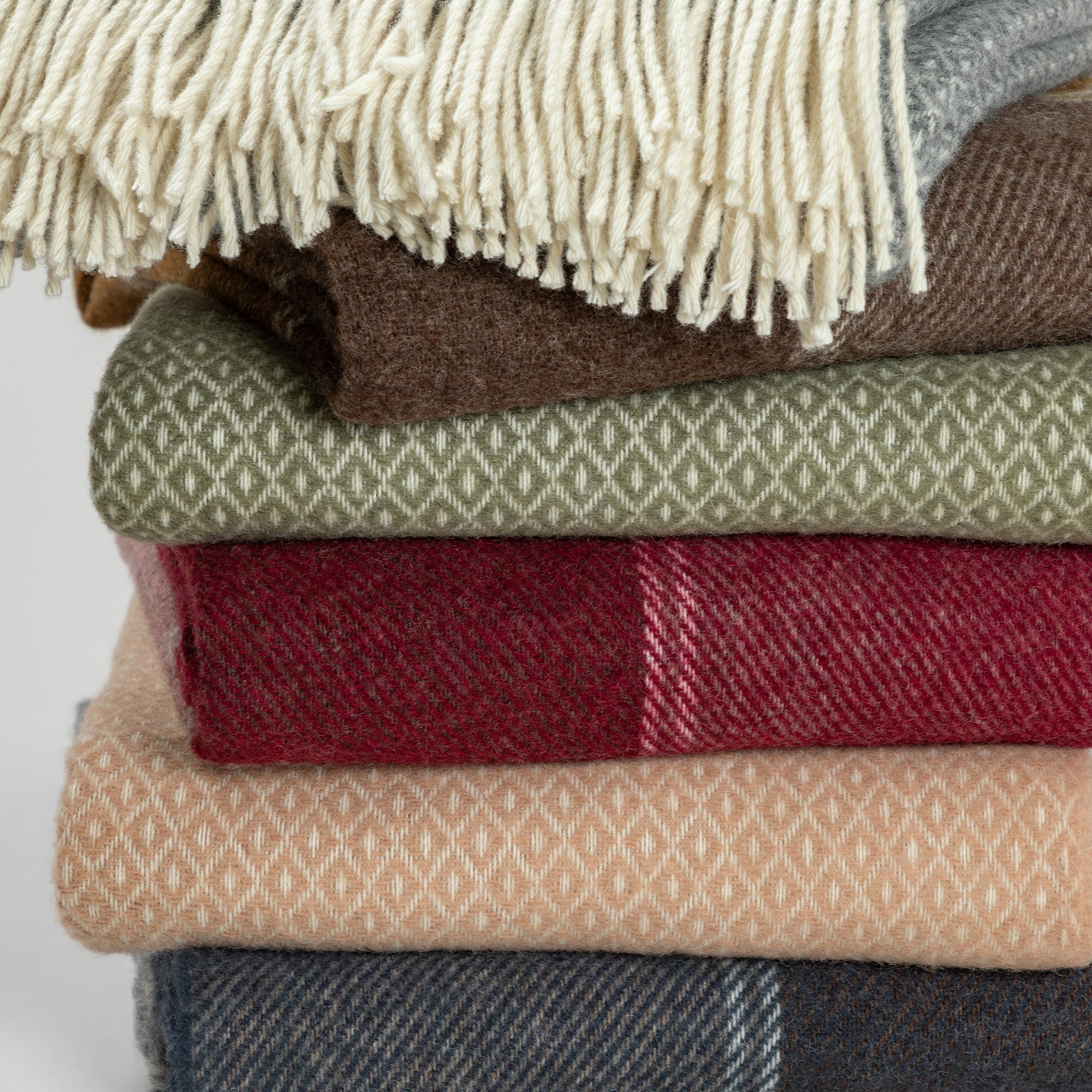 Close-up view of the stacked wool blankets focusing on the textures and fringes of the gray, brown, olive green, deep red, peach, and navy blankets.