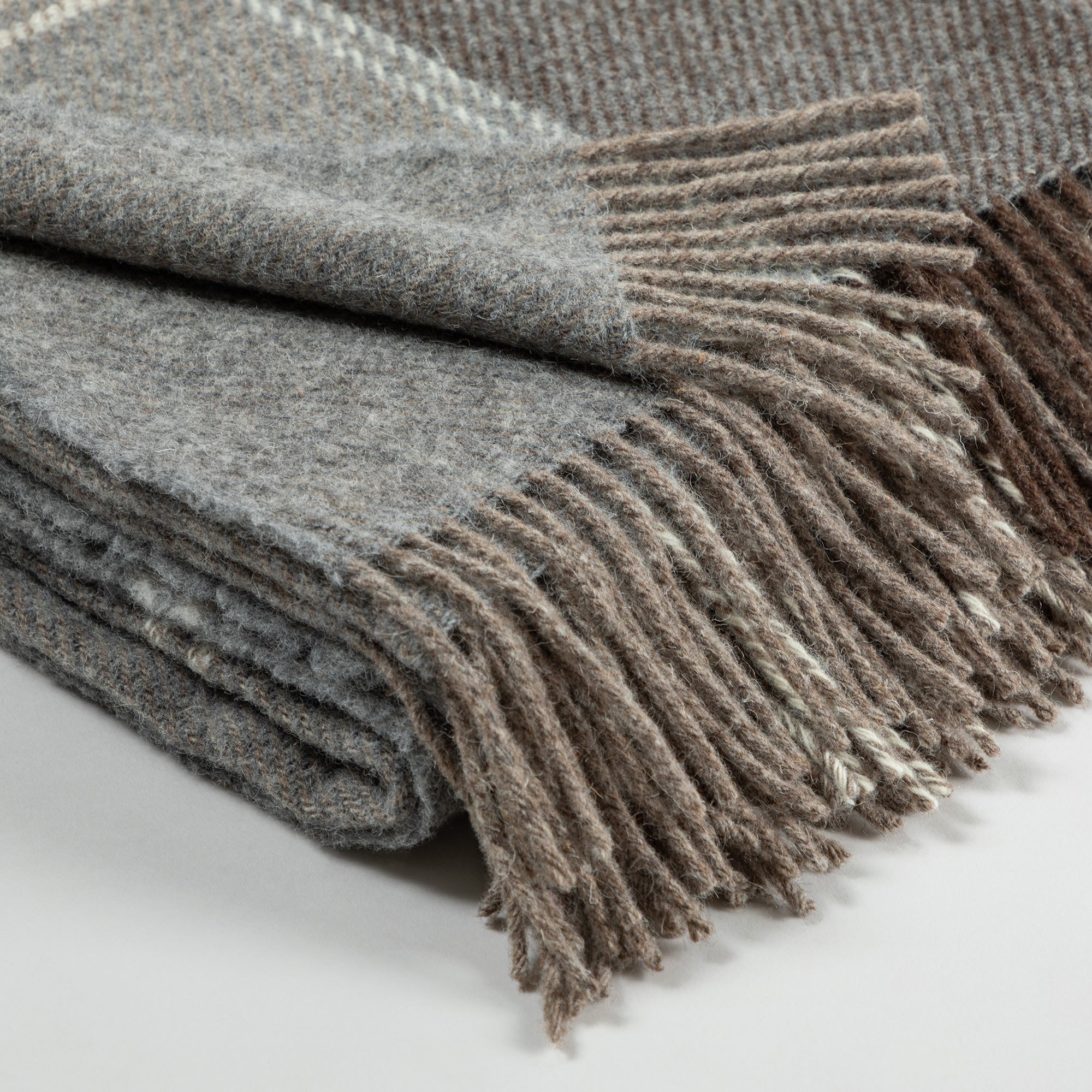 Close-up of a folded wool throw blanket with beige, brown, and gray plaid patterns and fringed edges on a neutral background.